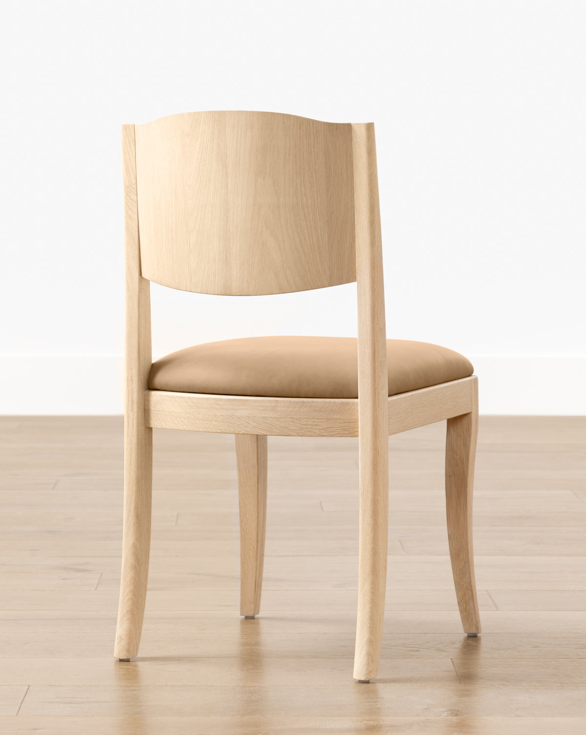 The Shepherd Dining Chair, featuring a white oak frame and tan upholstered seat cushion, is placed on a wooden floor against a plain white wall by McGee & Co.