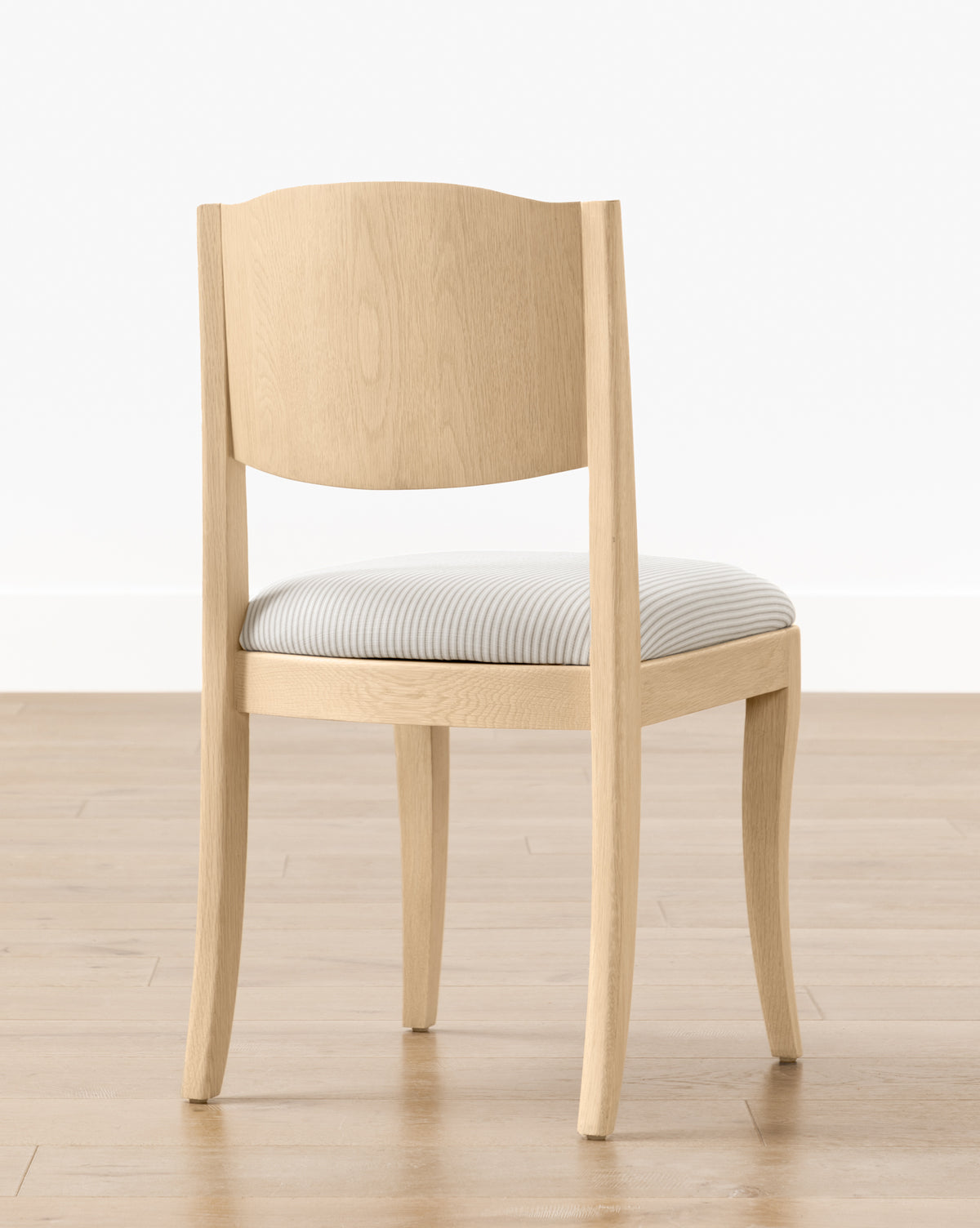 The Shepherd Dining Chair features light wood, a curved backrest, four legs, and a white and gray striped upholstered seat cushion. It is shown on a light wooden floor with a white wall background by McGee & Co.