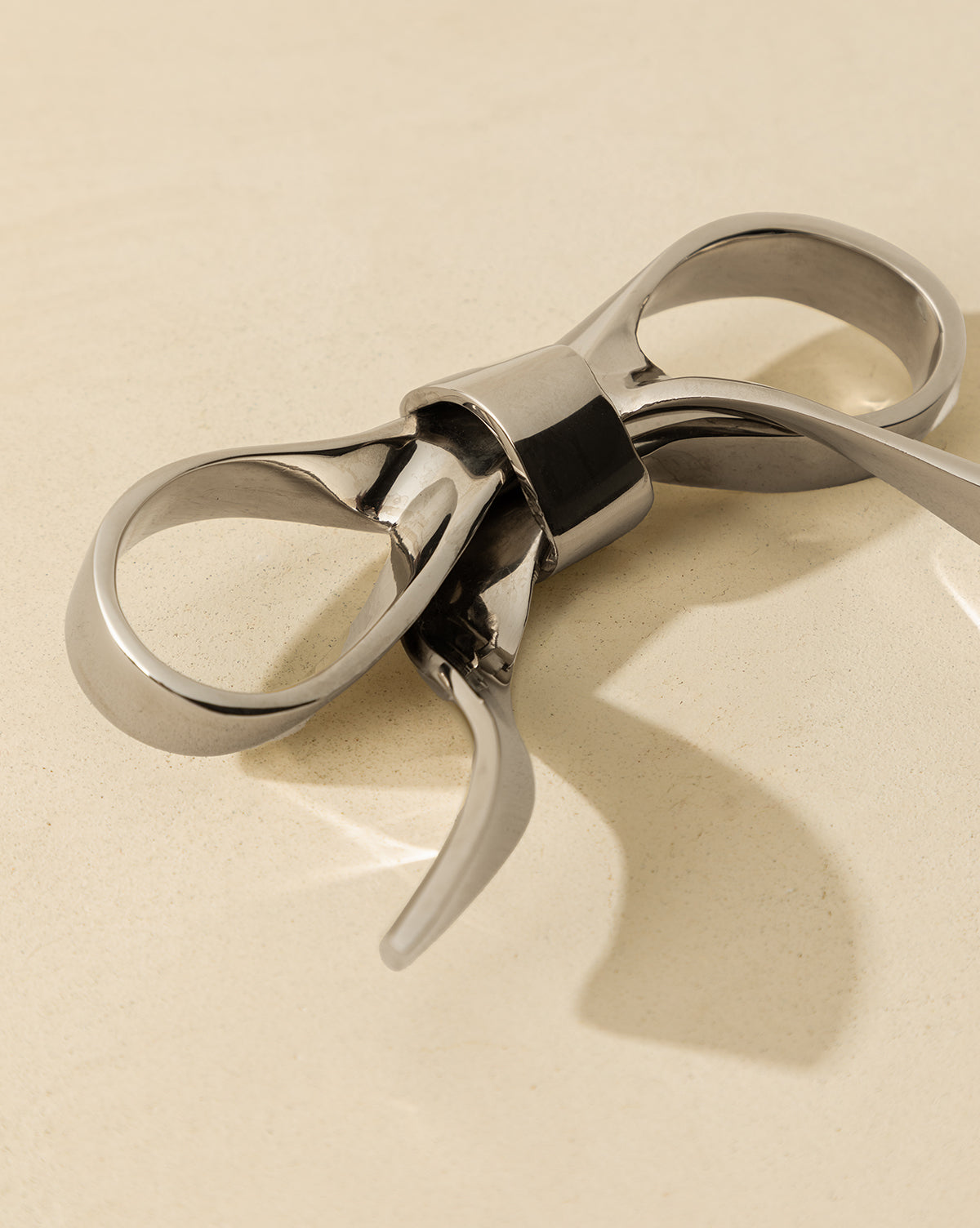 The Silver Bow Object, a lustrous silver decorative piece with elegant curved lines and vintage appeal, rests on a light beige surface, casting a gentle shadow, by Vendixs