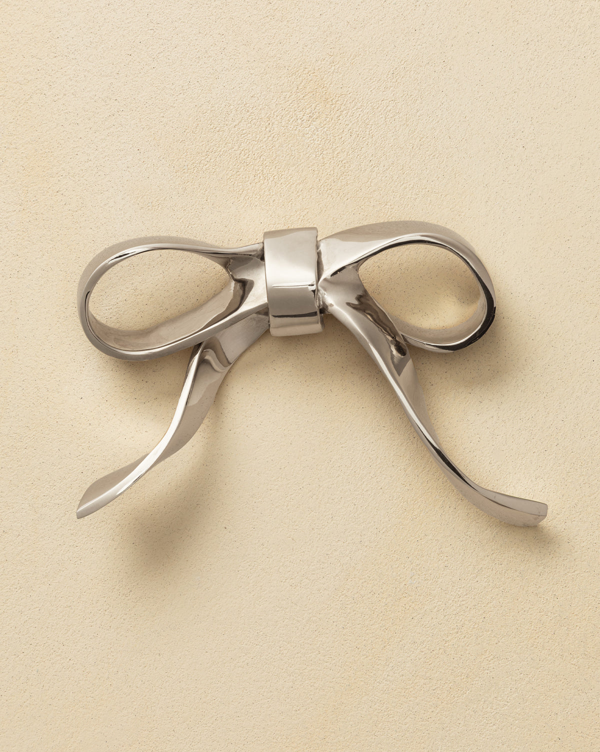 The Silver Bow Object is a shiny, metallic accent with smooth, curved loops and ends, displayed on a light beige, textured surface that radiates vintage charm, by Vendixs