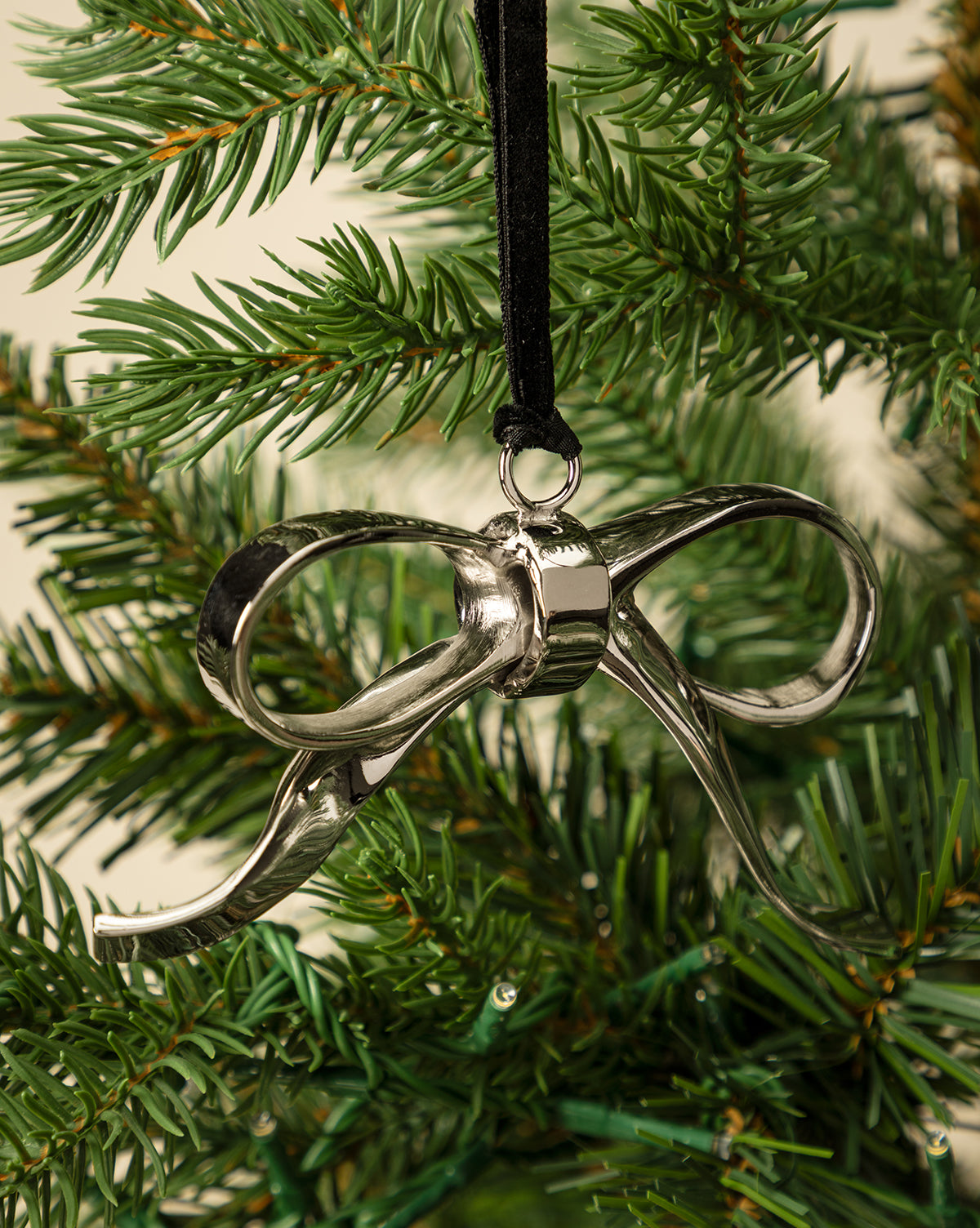 The Silver Bow Ornament, suspended from a black ribbon on a green holiday tree, brings festive charm and elegant décor to your seasonal celebrations, by Vendixs
