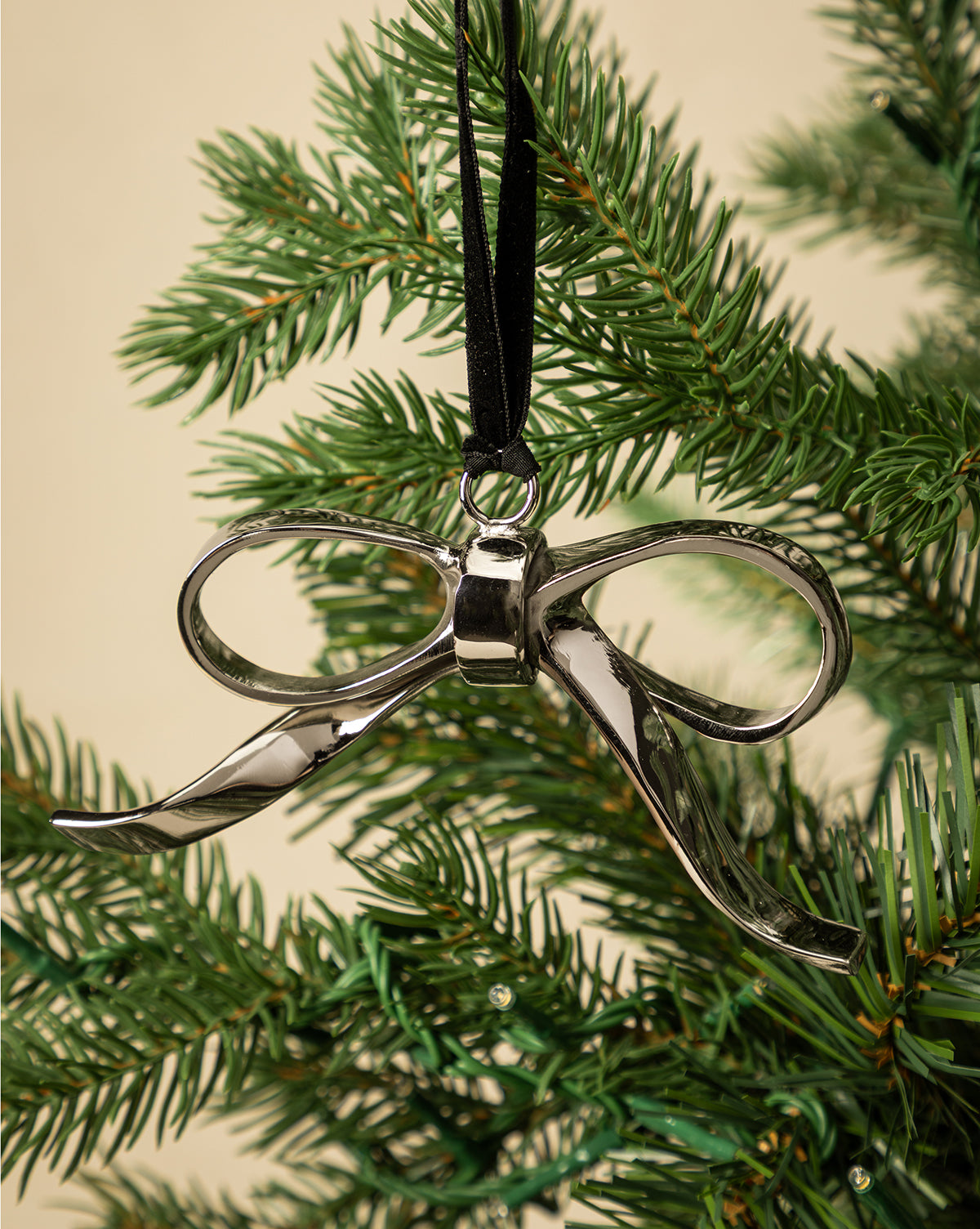 The Silver Bow Ornament hangs from a black ribbon on a green holiday tree, creating festive décor against a plain beige background, by Vendixs