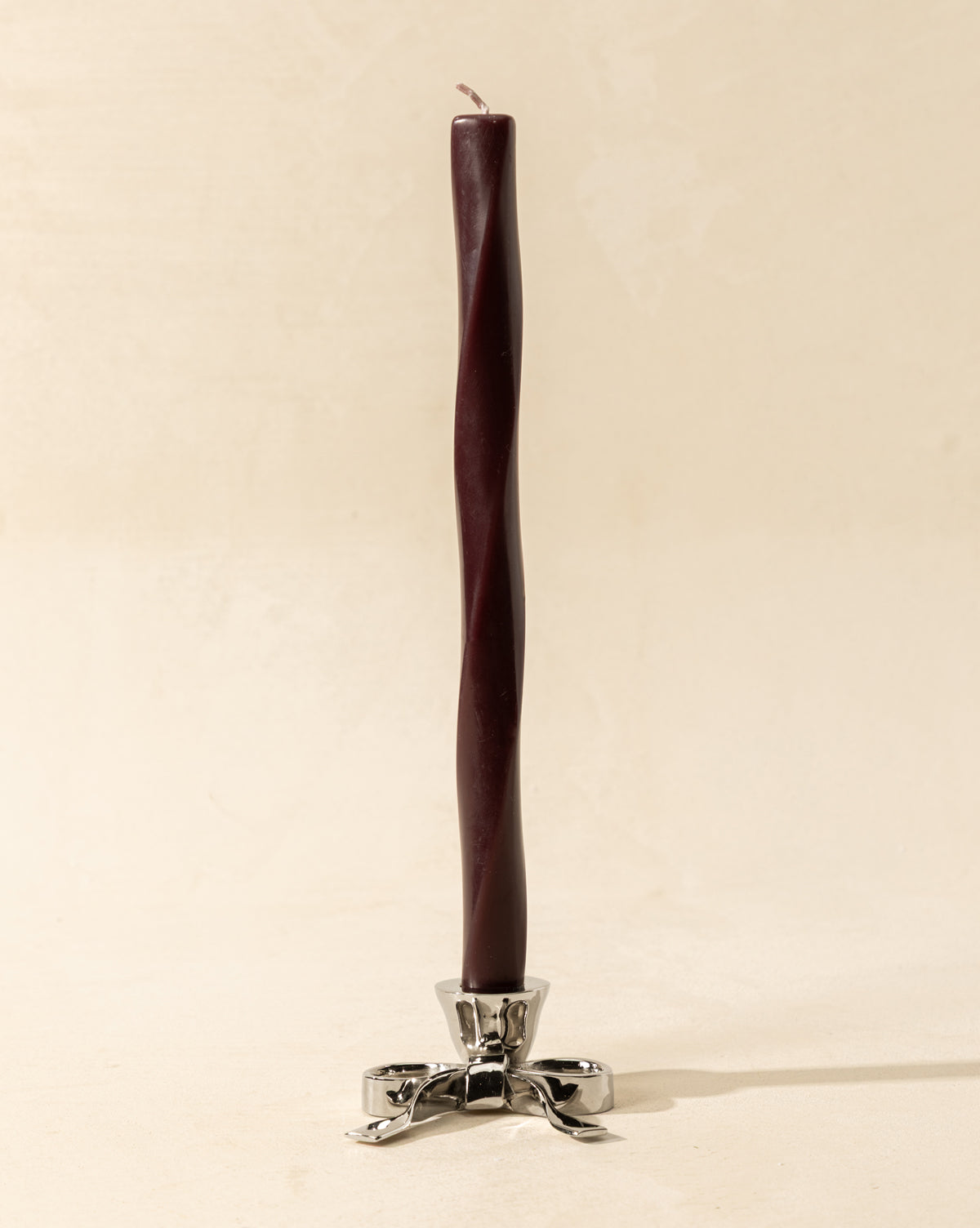 A tall, dark purple twisted taper candle stands in the Silver Bow Taper Holder, featuring three curved legs, making a striking accent for holiday tablescapes on a light beige surface, by Vendixs