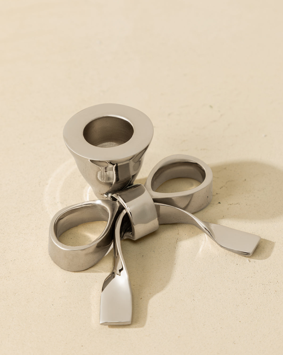The Silver Bow Taper Holder features a modern, metallic design with three rounded loops and paddle-like extensions—ideal for adding a sculptural touch to your holiday tablescape on a light beige surface, by Vendixs