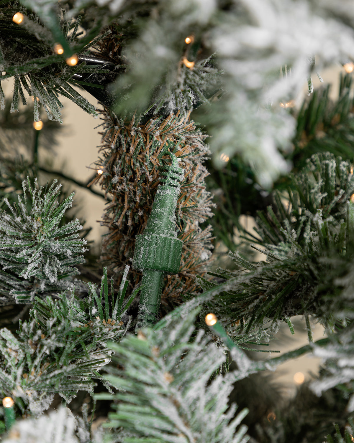 A close-up of the 7' Snowy Grandis Fir Pre-Lit Faux Tree displays a green electrical connector nestled among snow-dusted needles and glowing string lights on this realistic artificial Christmas tree, by Vendixs