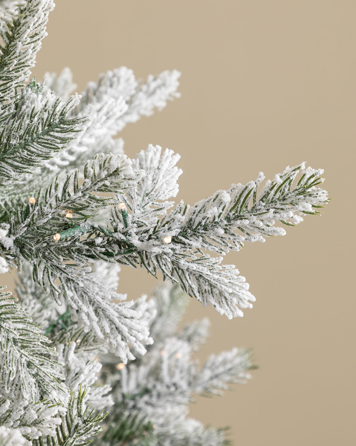 A close-up of snow-frosted branches from the 7' Snowy Grandis Fir Pre-Lit Faux Tree, adorned with small white lights, set against a plain beige background, by Vendixs