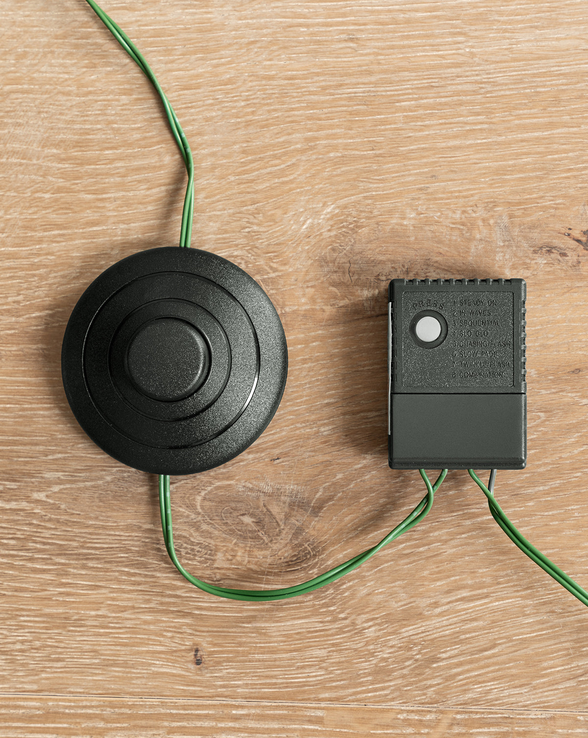 A round black wired foot pedal switch and a small rectangular control box with green wires from the 7' Snowy Grandis Fir Pre-Lit Faux Tree are both placed on a light wood surface, by Vendixs