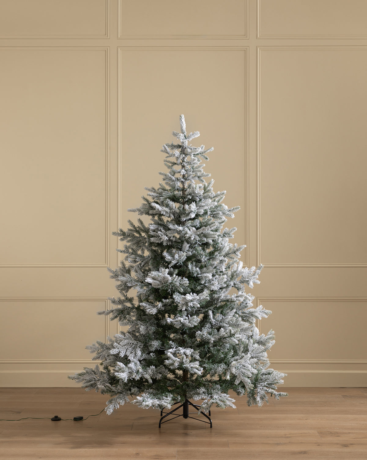 The 7' Snowy Grandis Fir Pre-Lit Faux Tree stands on a wooden floor by a beige paneled wall, with its unplugged power cord visible at the base, by Vendixs