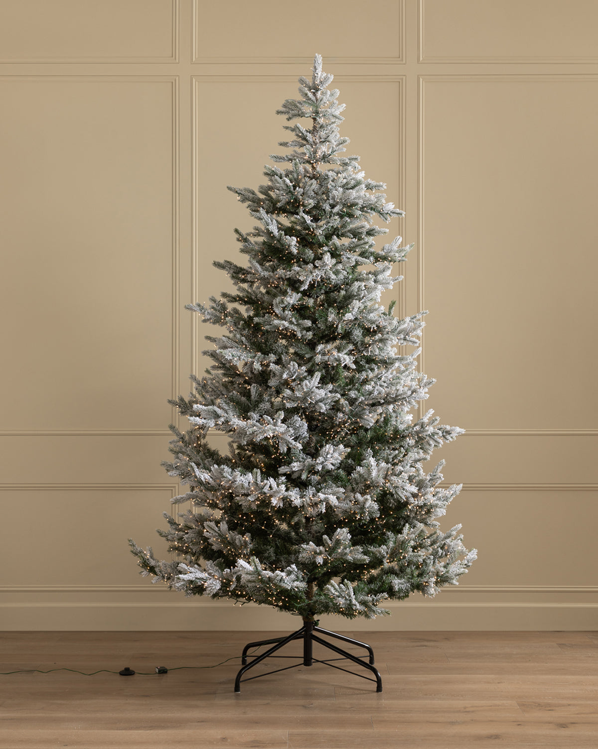The 9' Snowy Grandis Fir Pre-Lit Faux Tree stands on a black metal stand in a room with beige paneled walls and a wooden floor, featuring snow-frosted branches and small white lights, by Vendixs