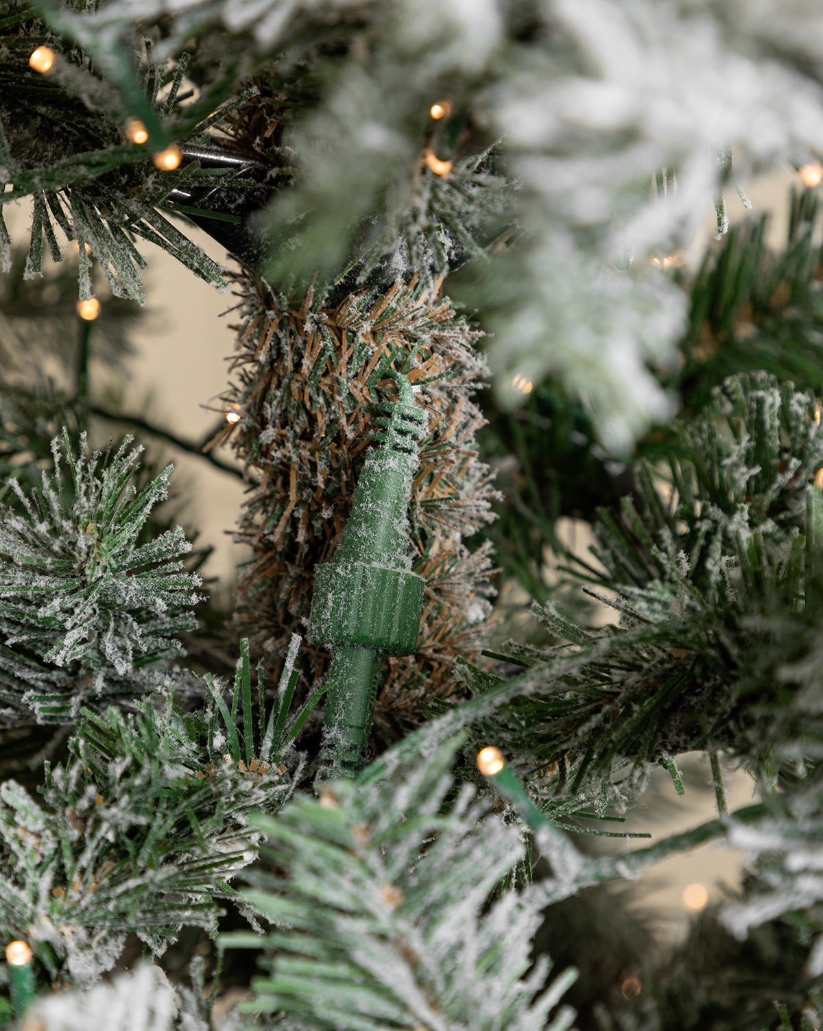 A close-up of the 9' Snowy Grandis Fir Pre-Lit Faux Tree shows a snow-dusted branch with a green electrical plug nestled in the needles and glowing white string lights in the background, by McGee & Co.
