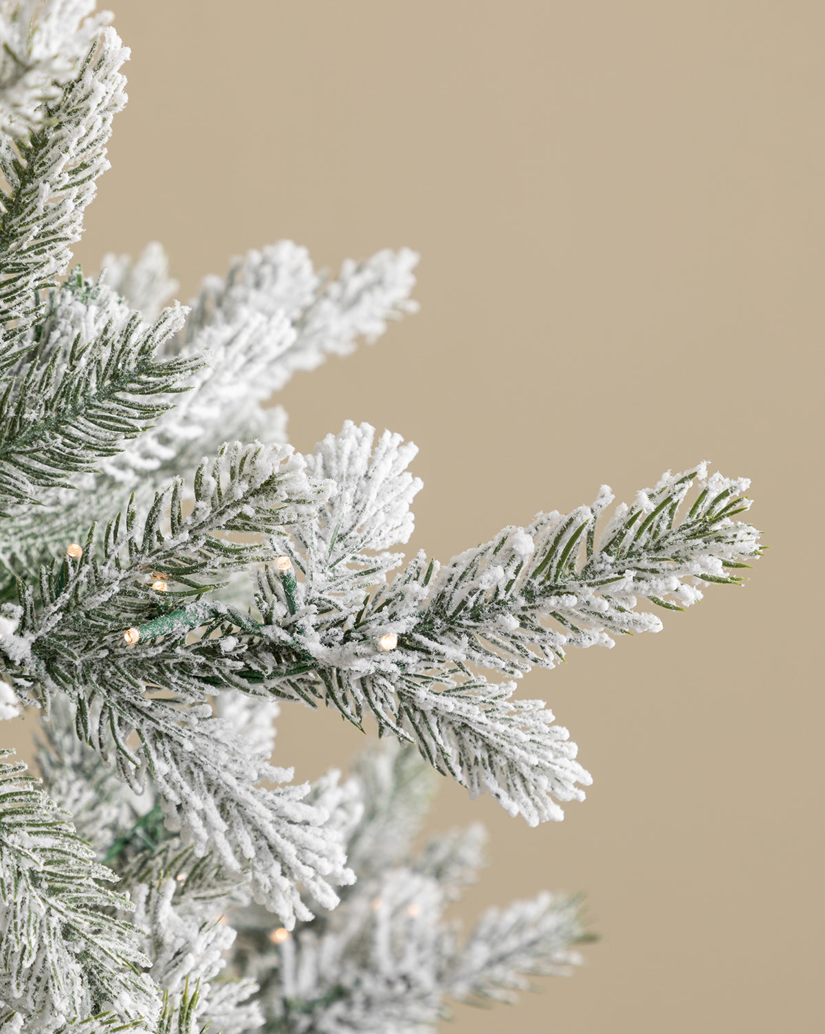 A close-up of the 9' Snowy Grandis Fir Pre-Lit Faux Tree shows green pine branches with white snow-like flocking and small white lights, set against a plain beige background, by Vendixs