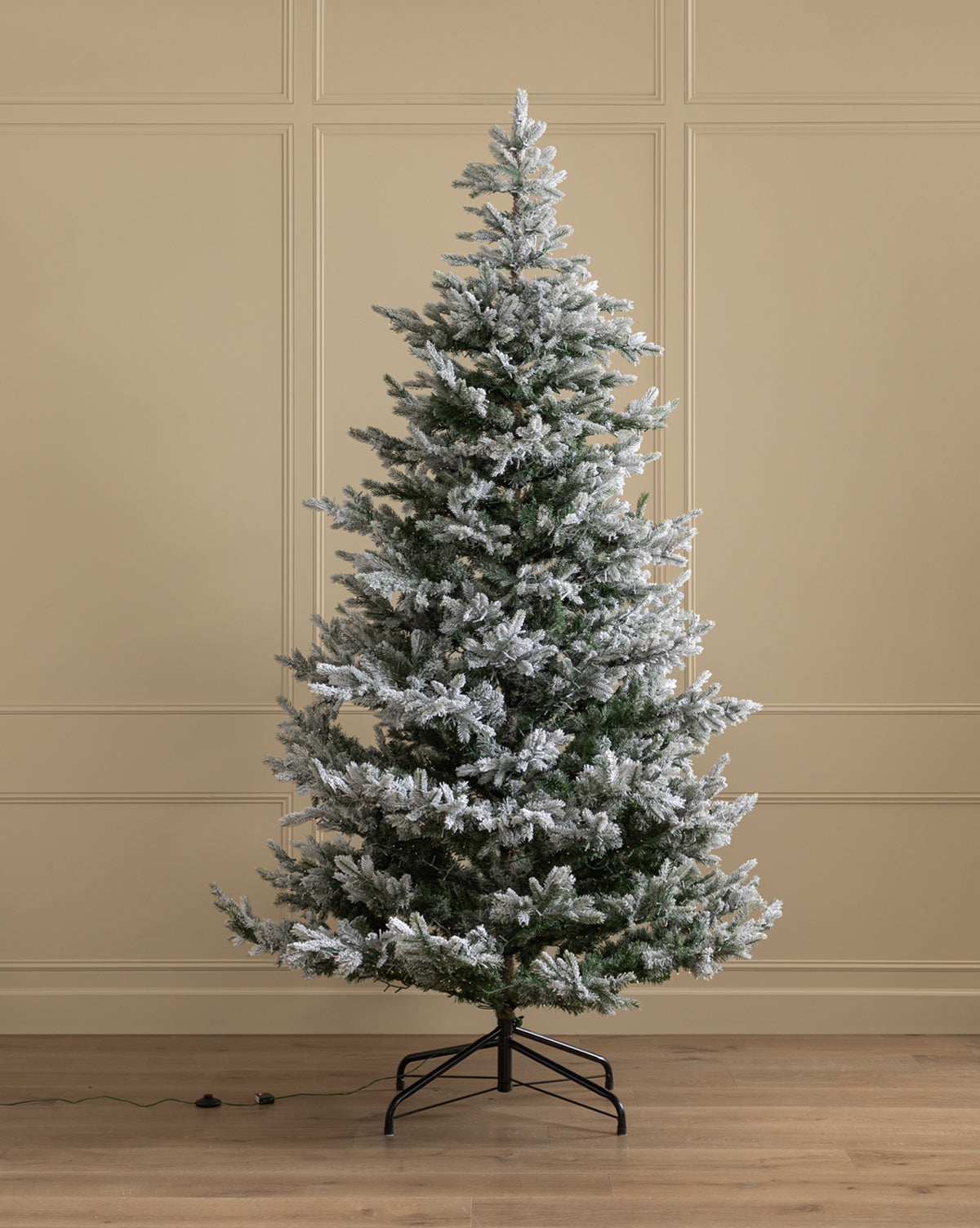A 9' Snowy Grandis Fir Pre-Lit Faux Tree stands on a black metal stand in a room with beige paneled walls and a wooden floor, with an electrical cord visible near the tree’s base, by Vendixs