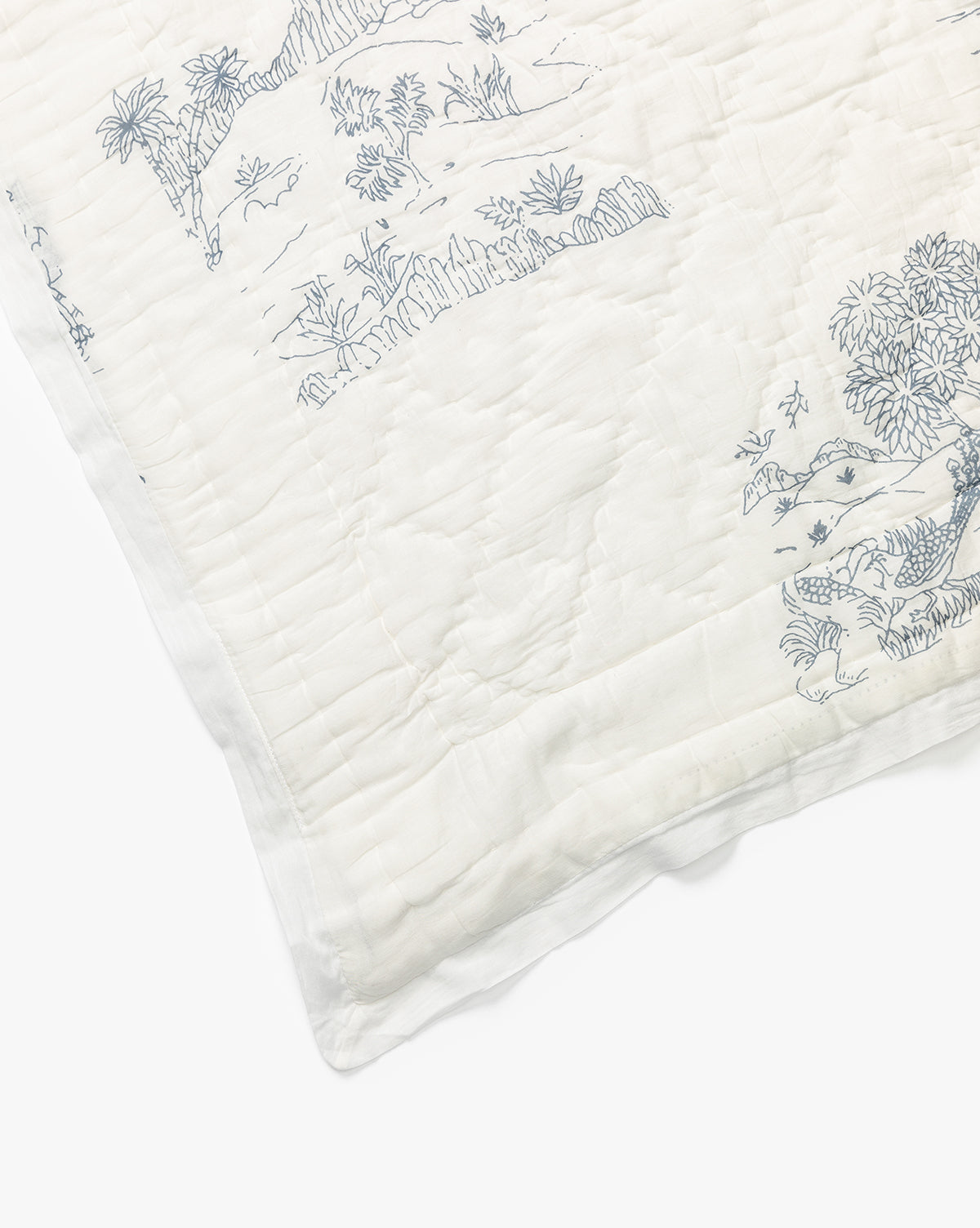 A corner of the Solano Block Print Kids Quilt by Indaba shows white cotton bedding with delicate blue line drawings inspired by nature scenes—trees, plants, and animals—creating a soft, charming look for kids. Vendixs