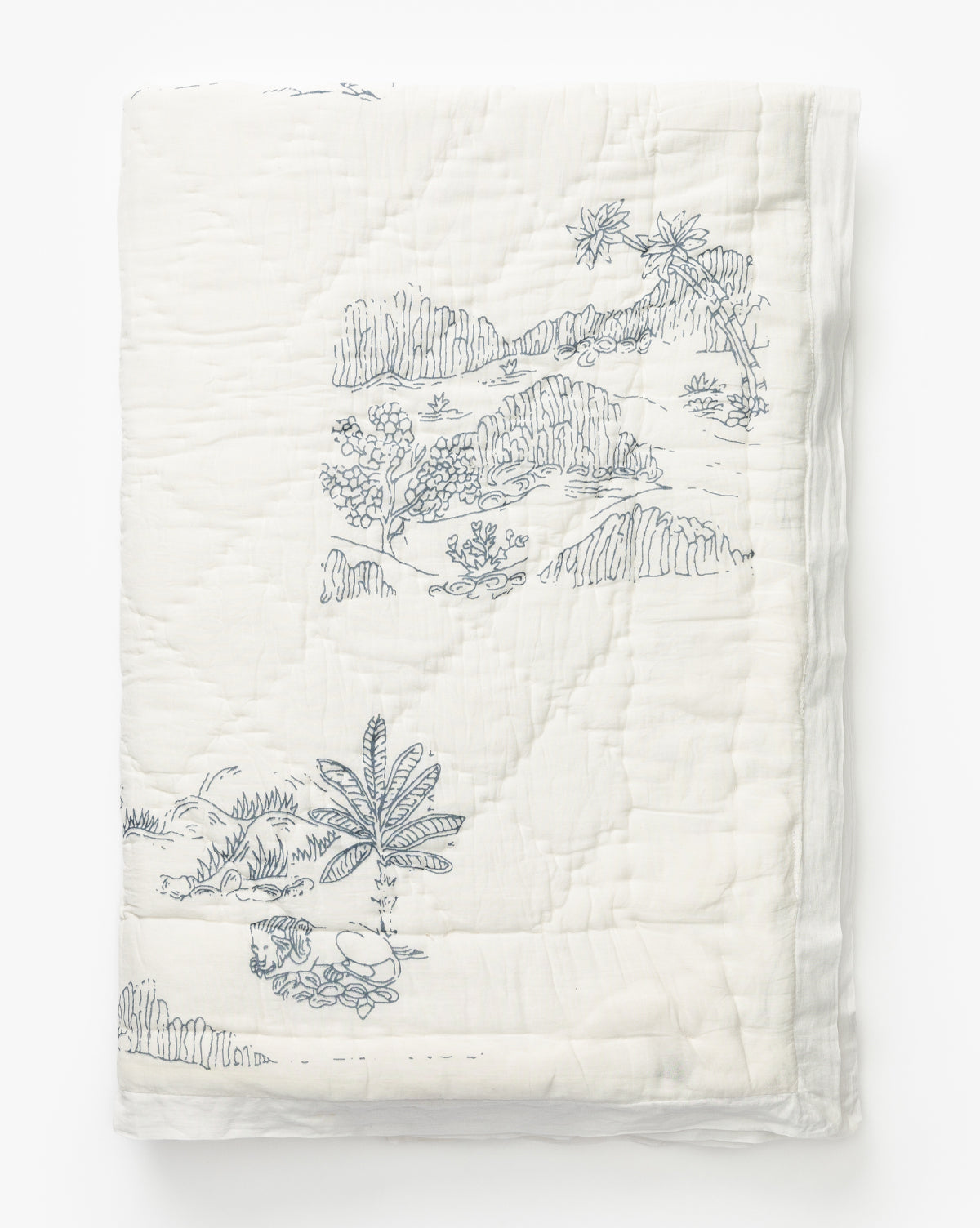 The Solano Block Print Kids Quilt by Indaba is a white cotton kids quilt featuring blue block print-style illustrations of landscapes with trees, hills, and a small dog, shown neatly folded against a plain white background. Vendixs