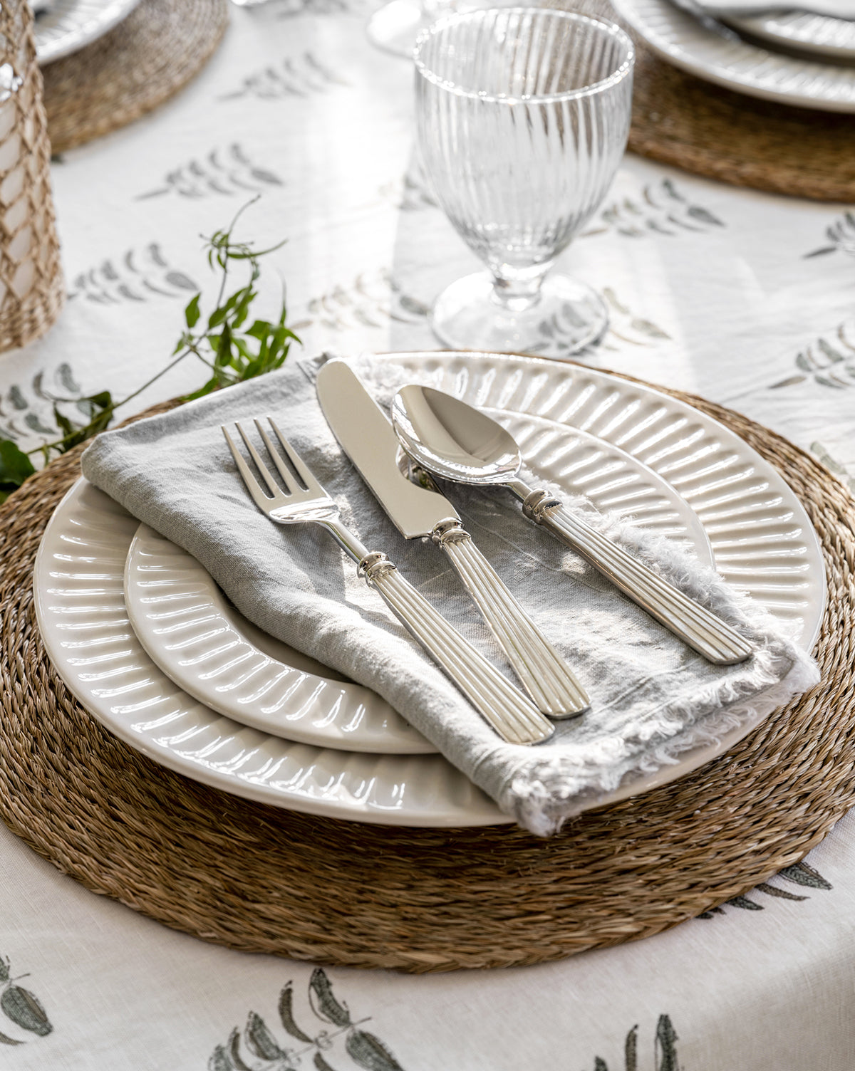 A close-up of a table setting with two stacked white plates, a gray napkin, the McNully Flatware (Set of 5) with textured handles, a clear glass, and a woven placemat on a white green-leaf-patterned tablecloth by McGee & Co.