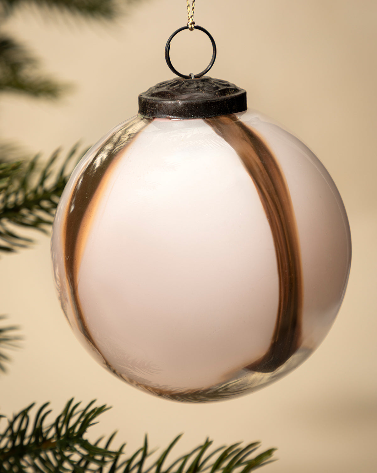 A close-up of the 4" Striped Glass Bauble in white and brown, hand-blown and hanging on a holiday tree branch with blurred green needles in the background, by Vendixs