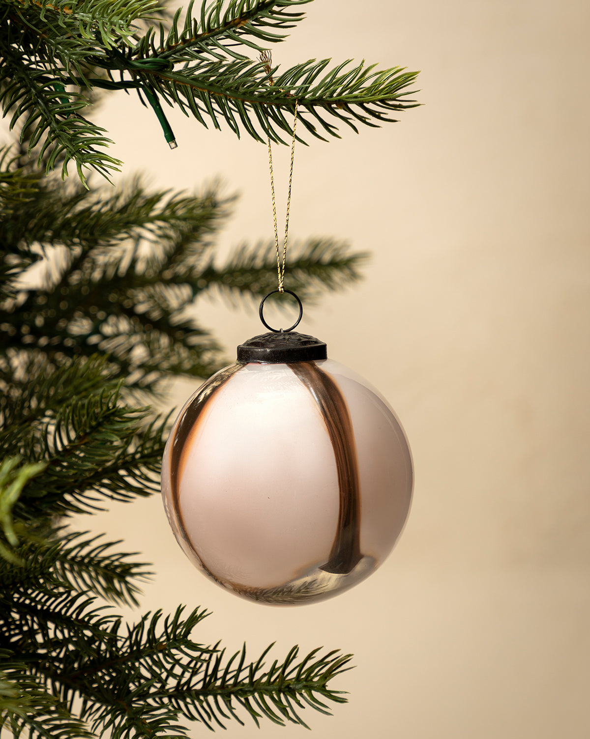 The 4" Striped Glass Bauble, crafted from hand-blown glass with dark vertical stripes, hangs from a gold string on a holiday tree branch against a plain beige background, by Vendixs