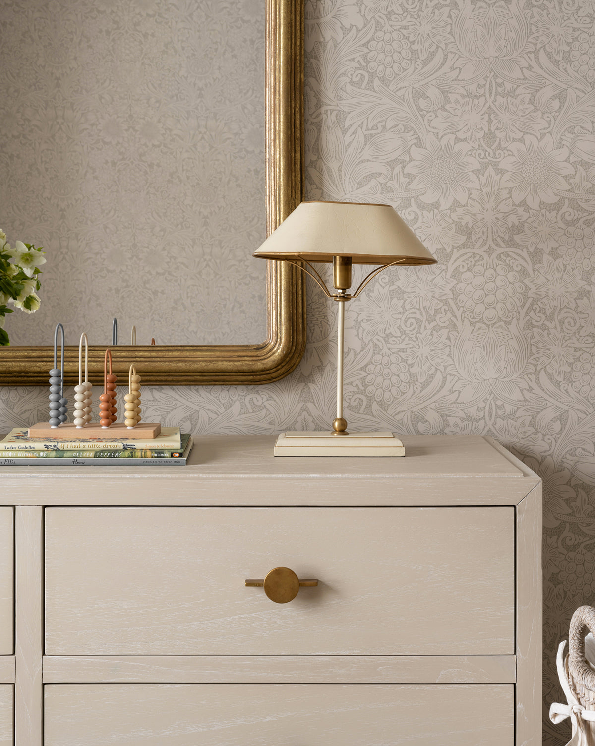 A beige dresser with a round gold knob stands against patterned wallpaper. On top, the Daphne Table Lamp with a beige shade sits next to a gold mirror, books, an abacus, art decor lamp, and a small green plant by Vendixs