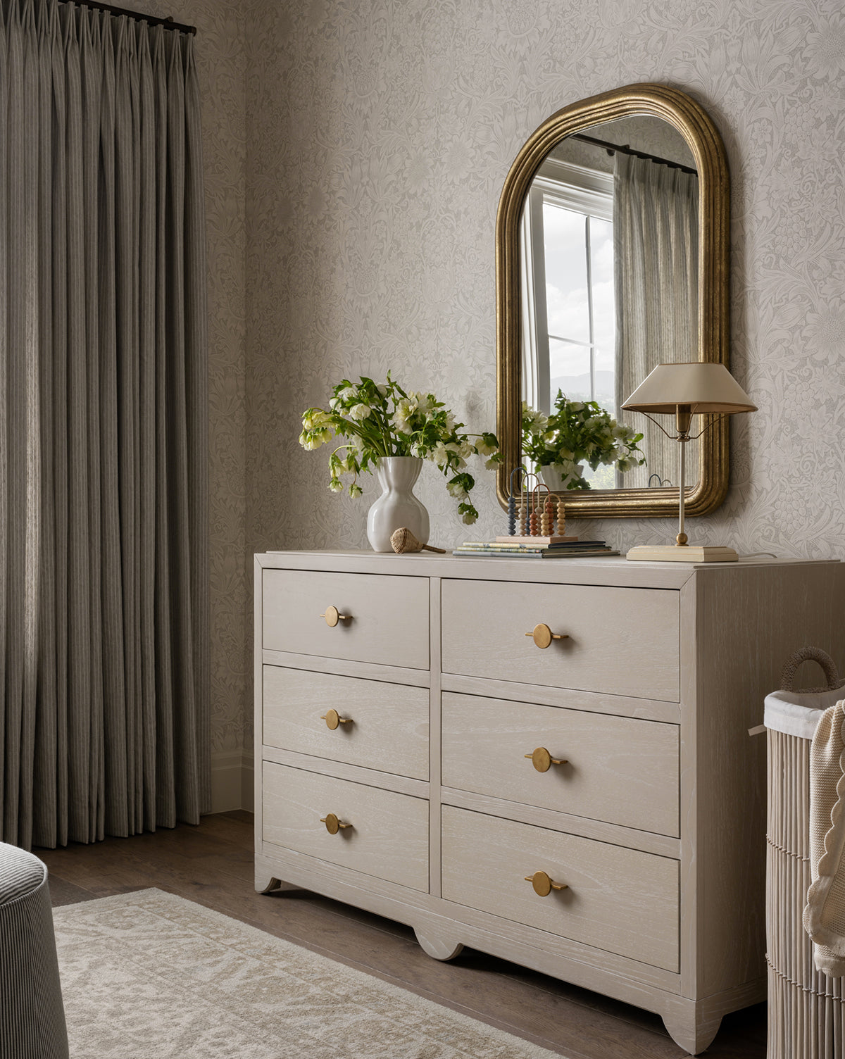 A light wood dresser with round gold knobs stands against patterned wallpaper. On top are a vase, lamp, books, and decor. Above it hangs the Giada Wall Mirror with an elegant moulded frame and antiqued finish, reflecting gray drapes nearby, by Vendixs