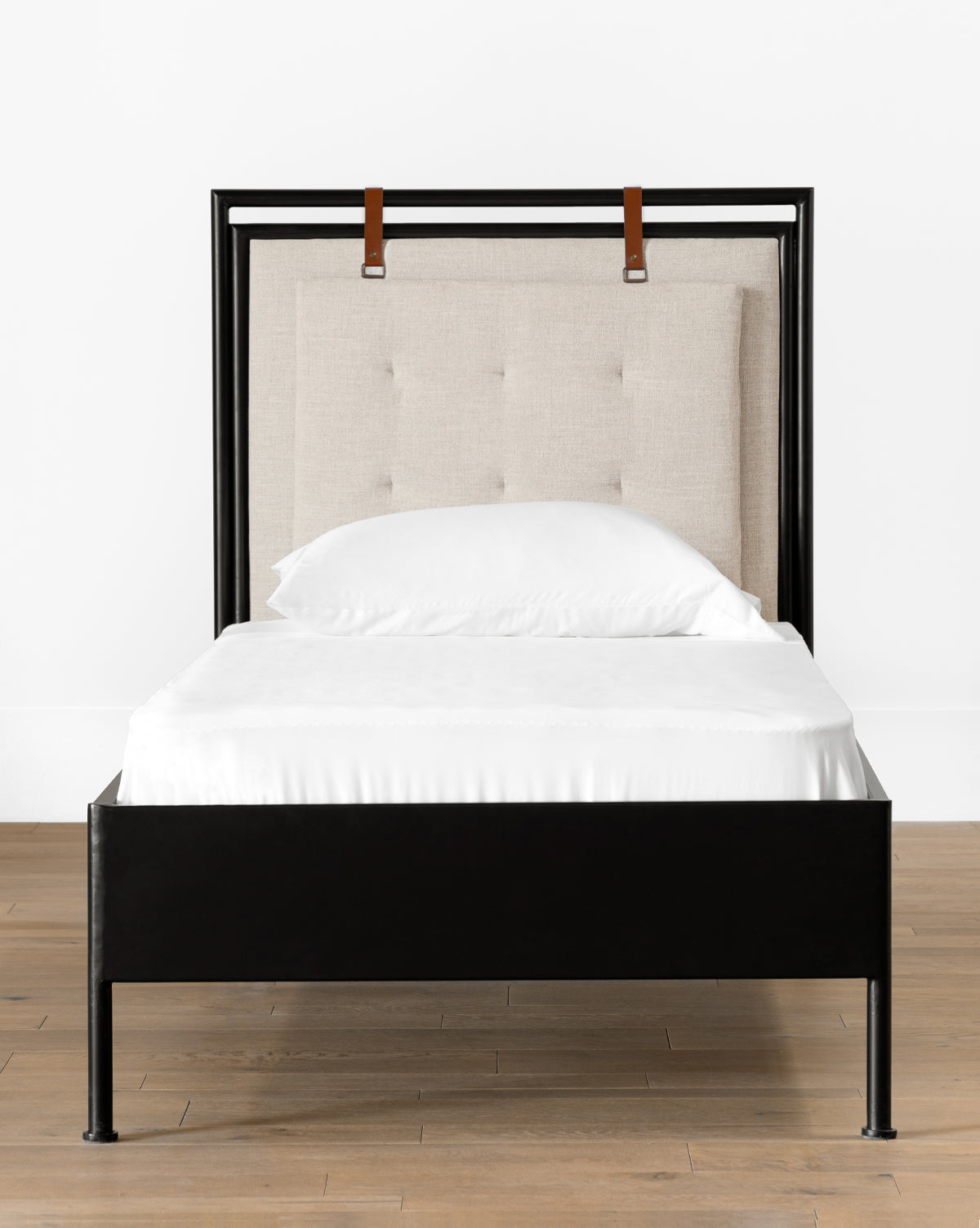 The Sutherland Twin Bed features a black metal frame, padded beige upholstered headboard with button tufting, and two brown leather straps at the top, set on a wooden floor against a white wall, by Vendixs