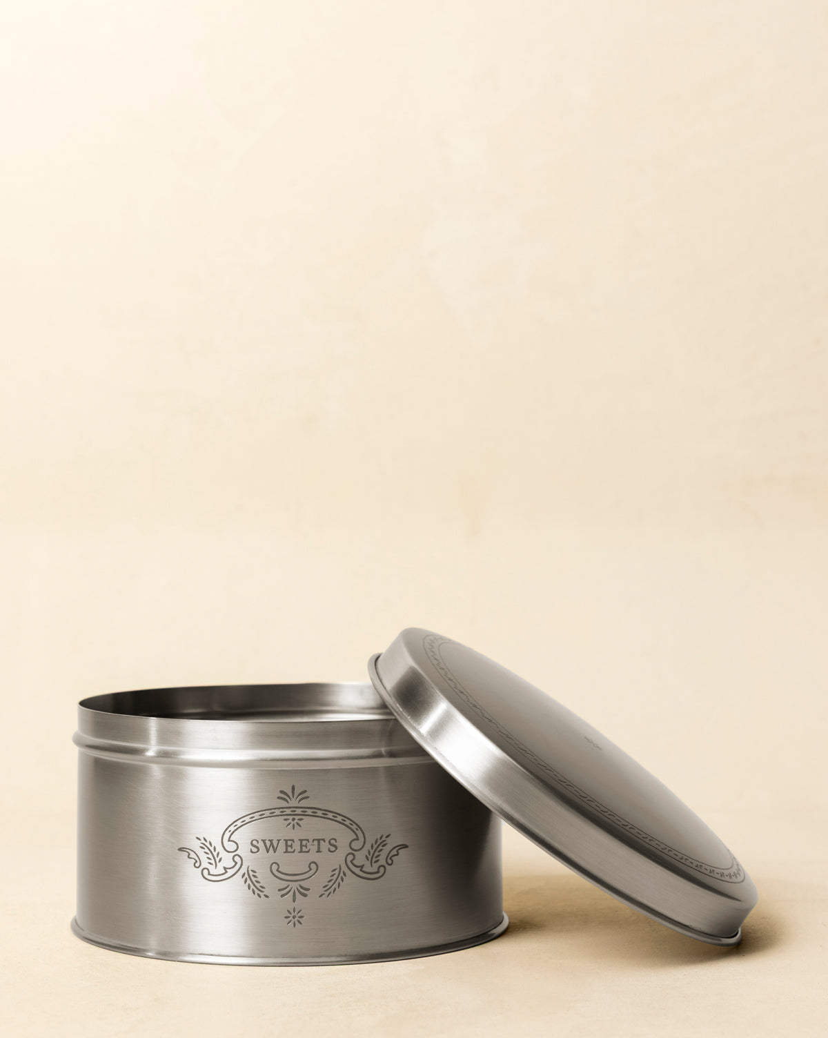 The Sweets Cookie Tin, a round silver metal container with a decorative label, sits open with its lid angled beside it, adding vintage charm against a plain beige background, by Vendixs