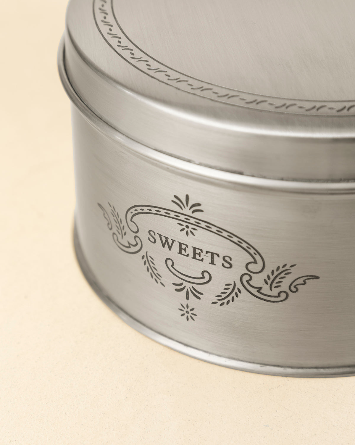 The Sweets Cookie Tin is a round, silver container with vintage-inspired decorative patterns and "SWEETS" engraved on the side, ideal for gifting holiday treats. It is displayed on a light beige surface, by Vendixs