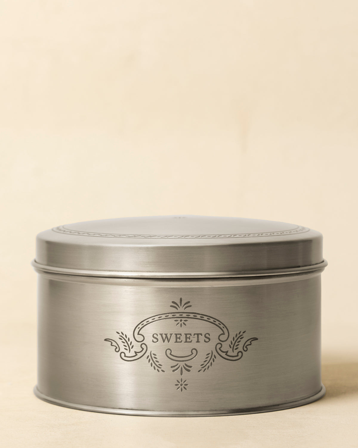 The Sweets Cookie Tin is a round, silver metal tin featuring a decorative pattern and "SWEETS" engraved on the front, adding vintage charm against a plain beige background, by Vendixs