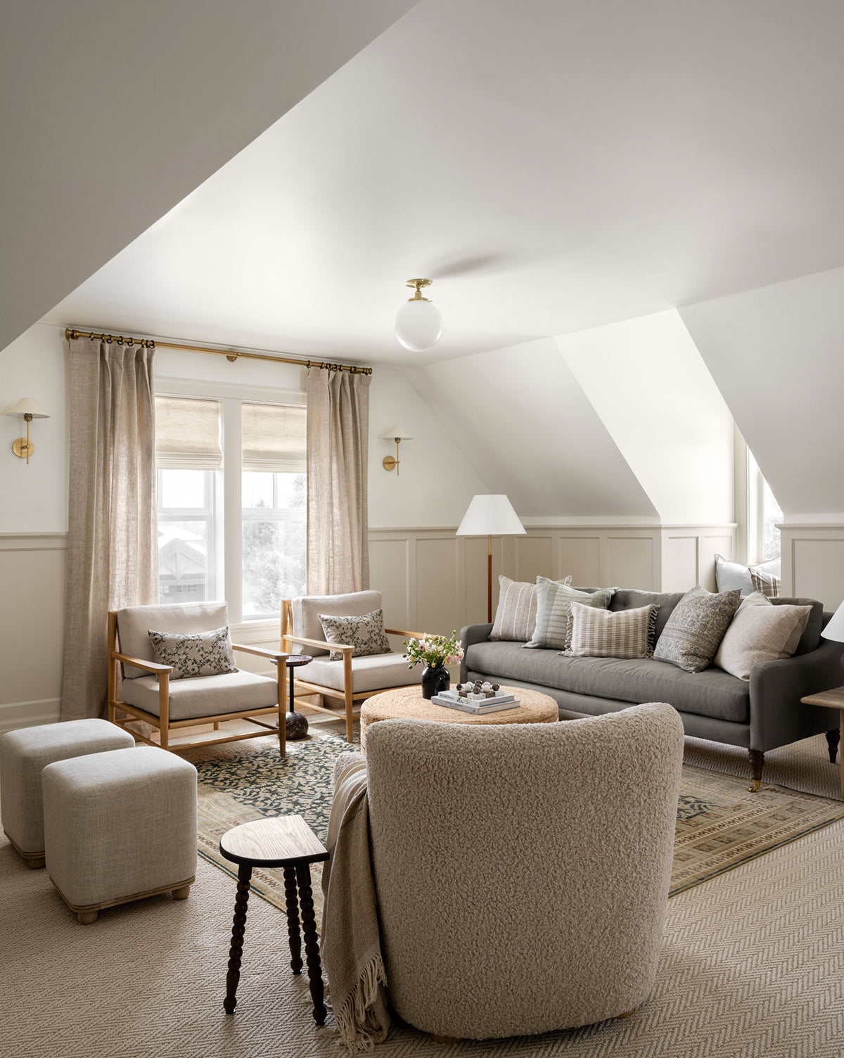 A cozy, neutral-toned living room with a gray sofa, light armchairs, soft rug, round table, ottomans, and an Alice Wood Stool by the large windows with beige curtains under sloped ceilings, by Vendixs