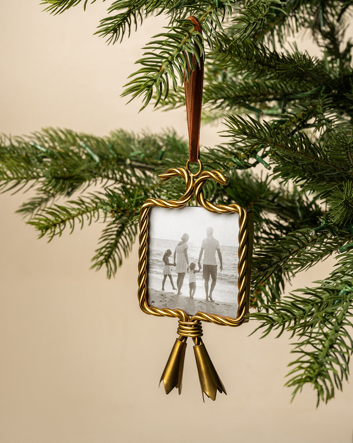 A Tassel Picture Frame Ornament with a gold frame, ribbon, and bells hangs on a Christmas tree branch. Inside is a black-and-white photo of a family of four holding hands, walking along the beach, by Vendixs