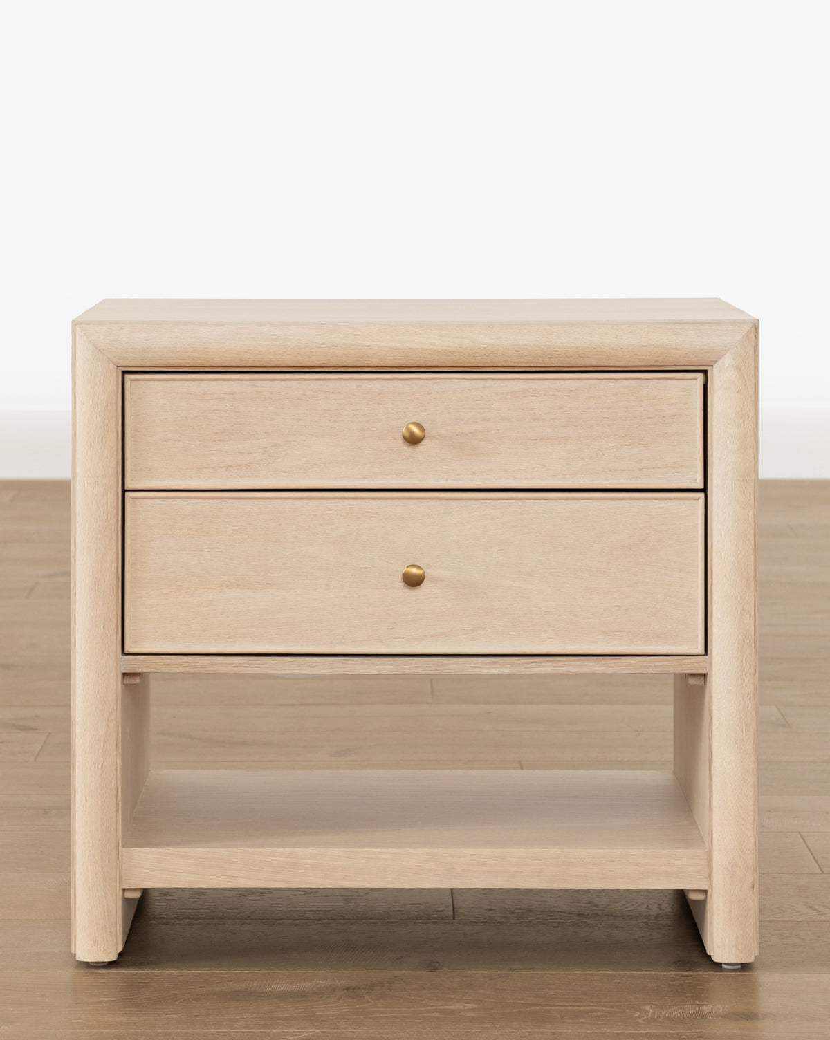 The Tellerbeck Nightstand showcases a light, bleached oak finish with two drawers accented by gold knobs and an open lower shelf, ideal for bedroom styling, by Vendixs Displayed on a wooden floor with a plain white backdrop.