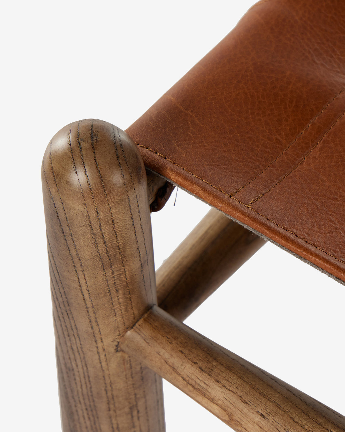 Close-up of the Tierney Ottoman by Four Hands, featuring a solid wood leg with natural grain and smooth finish, topped with a brown stitched top-grain leather seat that is slightly textured. Vendixs