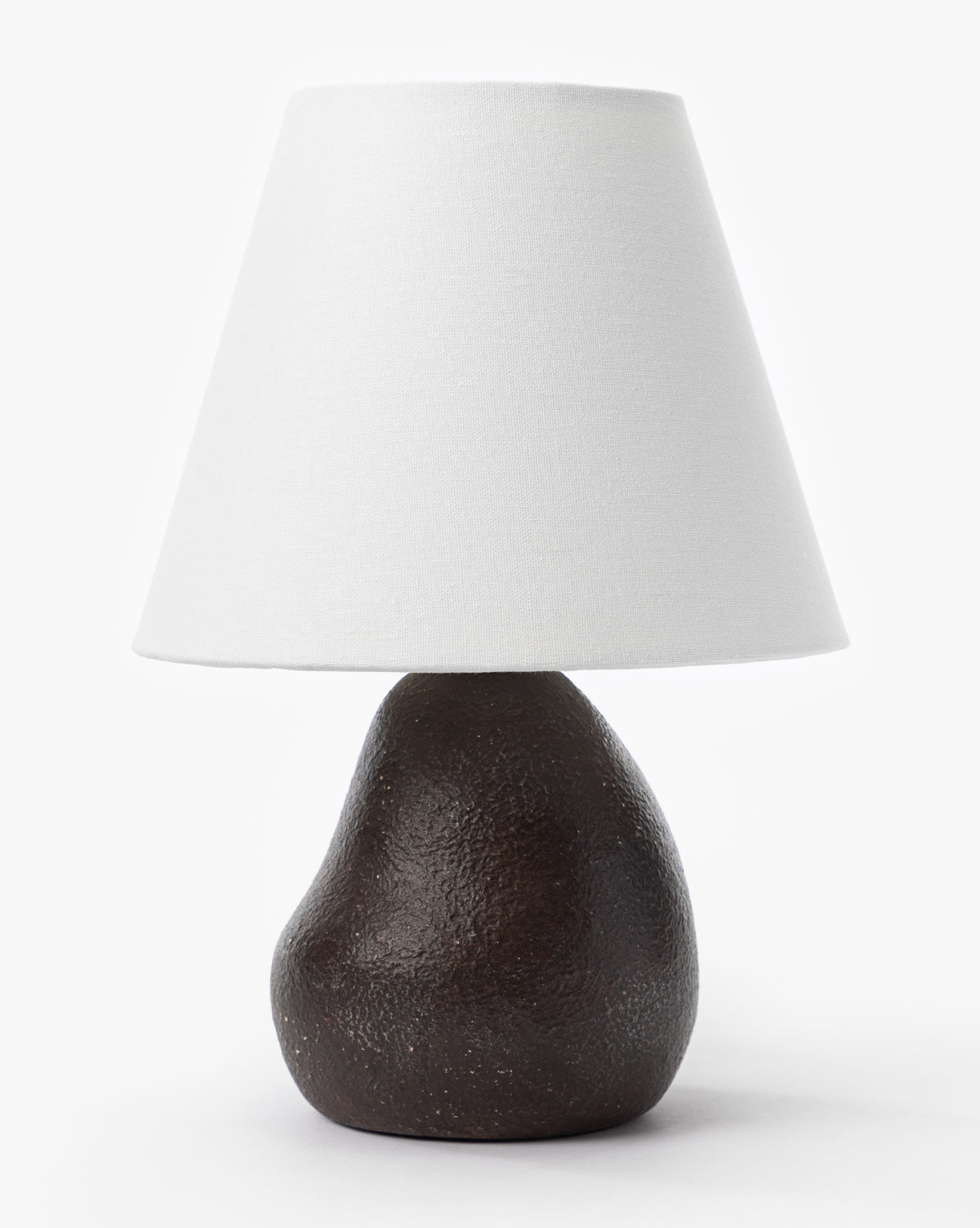 The Makerspalm Vedruna Table Lamp showcases a textured dark brown ceramic base with a large tapered white shade, all presented against a plain white background. Vendixs
