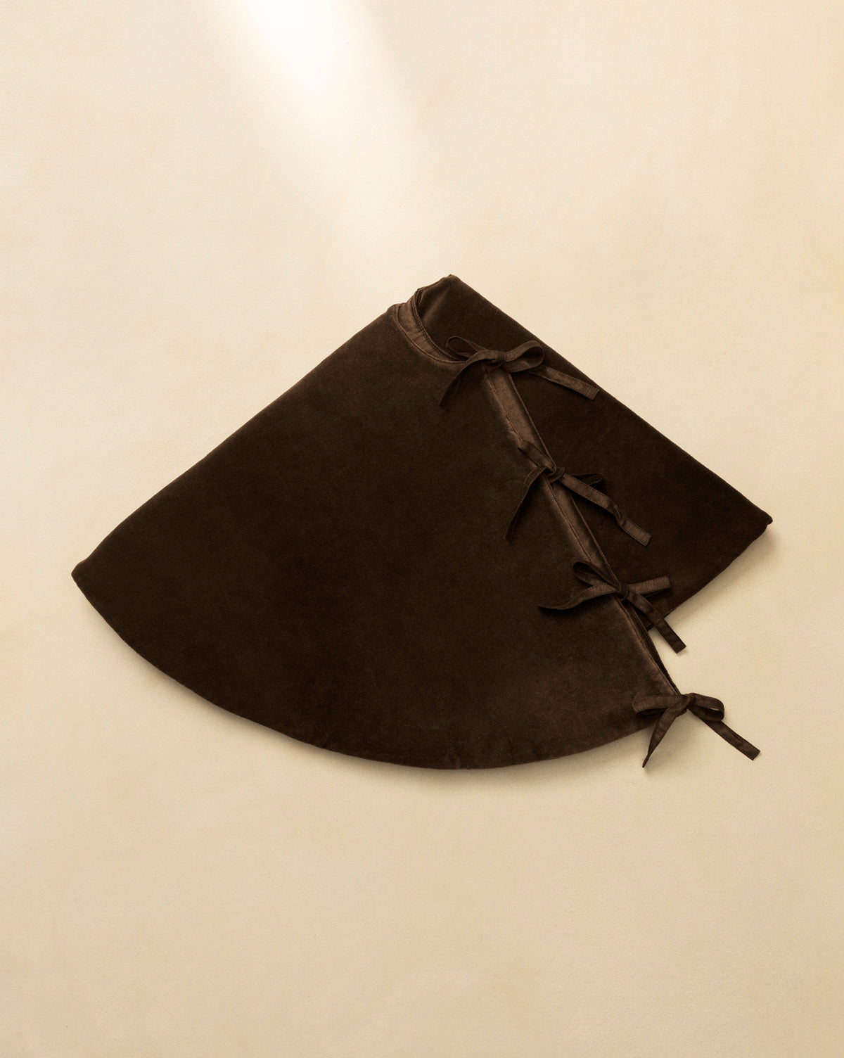 The Velvet Brown Tree Skirt, a dark brown semi-circular design with three ribbon tie closures, is displayed flat on a beige background, highlighting its luxurious texture—ideal for stylish Christmas tree decor, by Vendixs