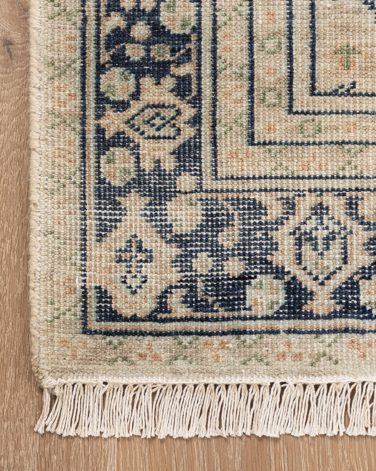 A close-up of the Vercelli Hand-Knotted Wool Rug corner shows its fringed edges and geometric floral patterns in navy blue, beige, and green on a light wooden floor, by Vendixs