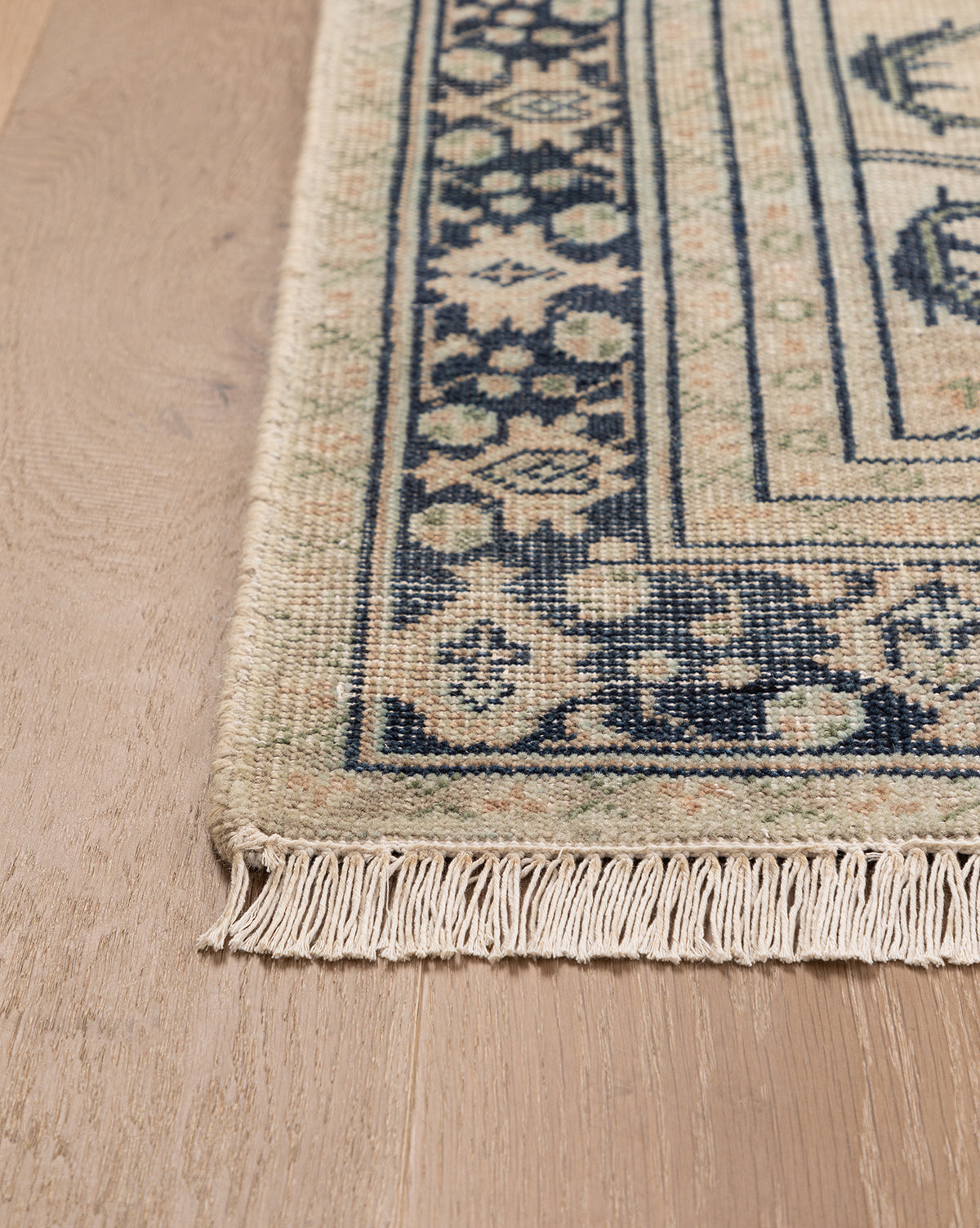 A close-up of the Vercelli Hand-Knotted Wool Rug with fringe, shown on a light wood floor. The rug displays beige and navy geometric and floral border motifs, by Vendixs