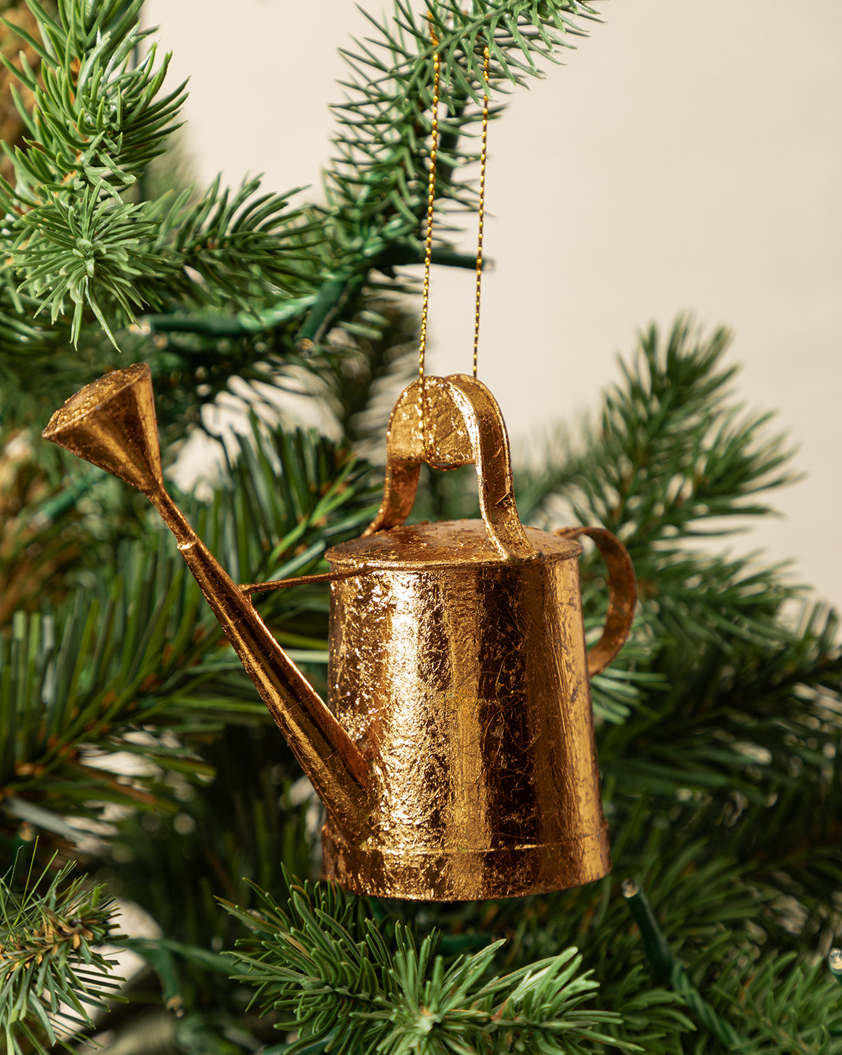 The Watering Can Ornament hangs from a green Christmas tree branch, adding unique charm to your festive decor with its copper finish, by Vendixs