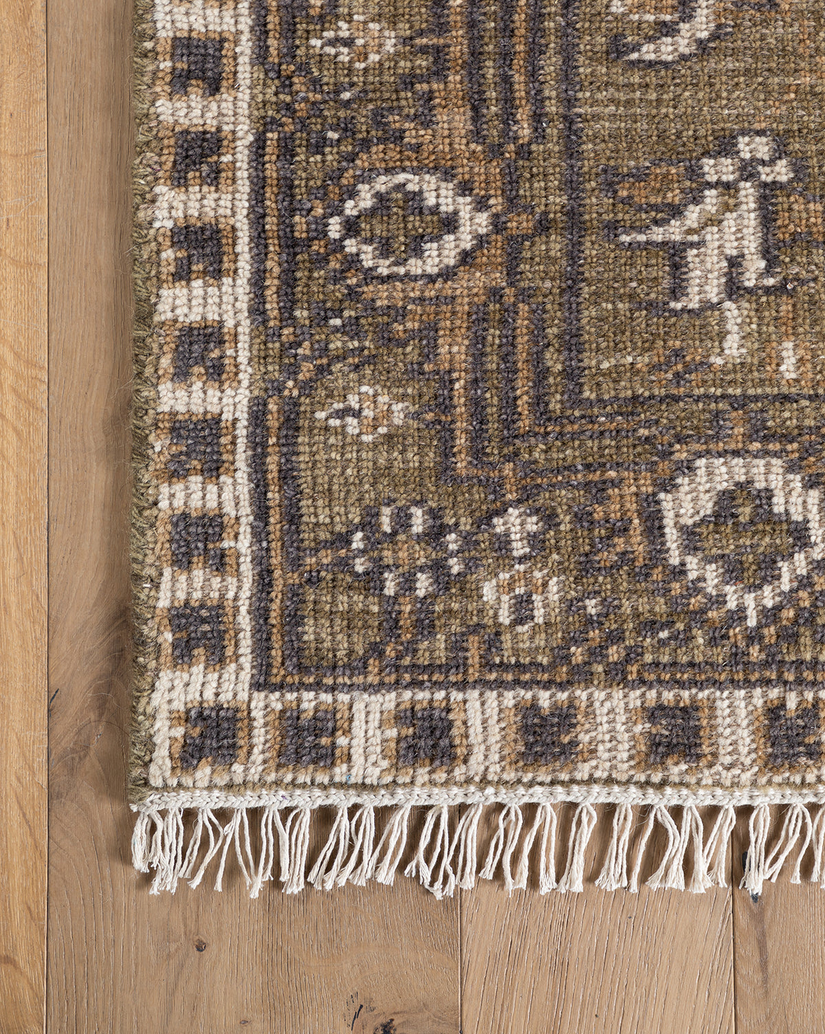 A close-up of the Wayland Hand-Knotted Wool Rug shows its brown, beige, and dark gray geometric and floral patterns with fringe along the edge, displayed on a light wooden floor, by Vendixs