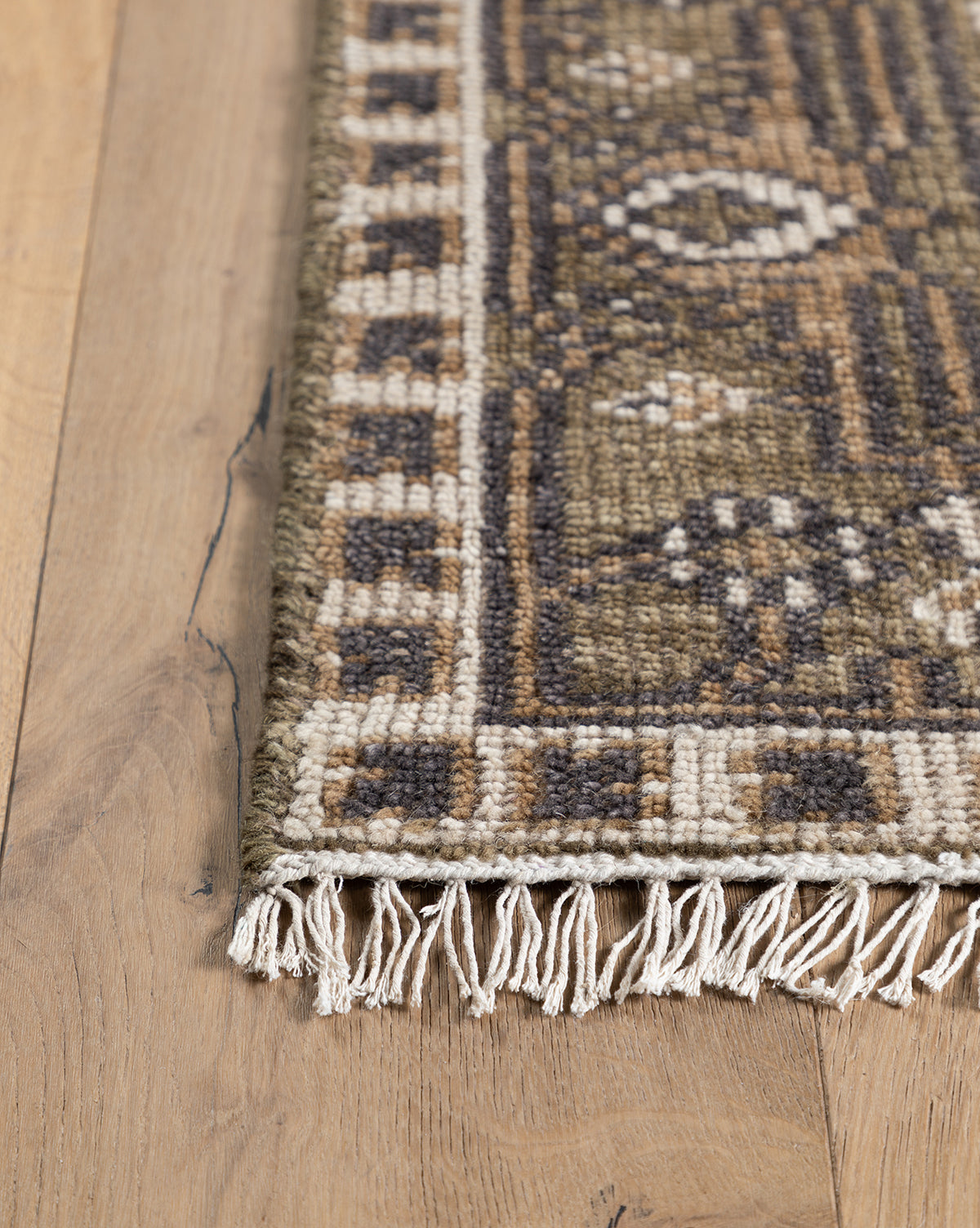 A close-up of the Wayland Hand-Knotted Wool Rug shows its decorative border and white fringe on a light wood floor. The artisan wool rug features earthy tones of brown, beige, and olive, by Vendixs