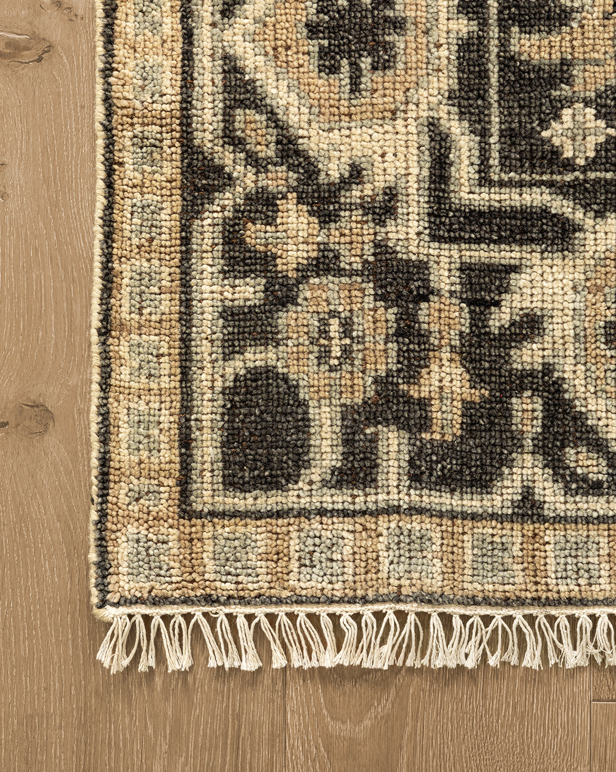 A close-up of the corner of a Wayland Hand-Knotted Wool Rug highlights its beige, black, and brown floral pattern with fringe, displayed on a light wood floor by Vendixs