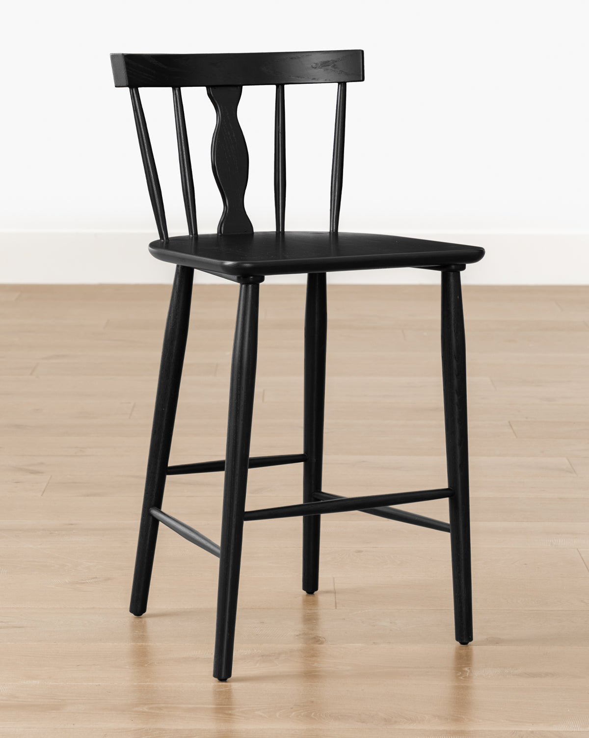The Whittaker Counter Stool by YANG CHENG WOODEN INDUSTRIES INTERNATIONAL (VIETNAM) CO LTD features a black wood finish with a curved spindle back, three vertical spindles, and a decorative center detail, shown on a light floor against a white wall. Vendixs