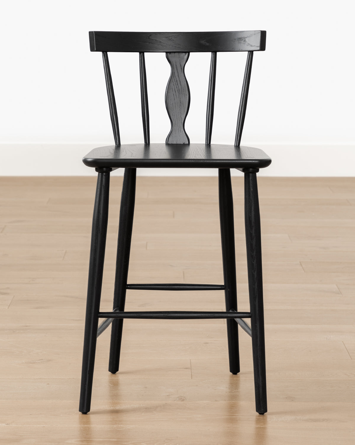 A black Whittaker Counter Stool by YANG CHENG WOODEN INDUSTRIES INTERNATIONAL (VIETNAM) CO LTD, featuring a spindle back and decorative center, crafted from solid oak, sits on a light wood floor against a plain white wall, Vendixs