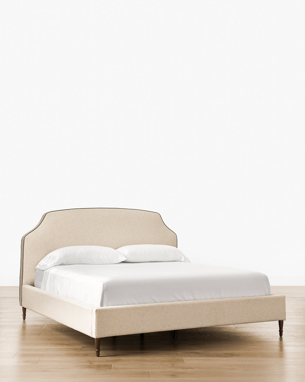 The Willoughby Bed showcases beige linen upholstery, a curved headboard, English Oak legs, white bedding with two pillows, set on wooden flooring against a plain white backdrop, by Vendixs