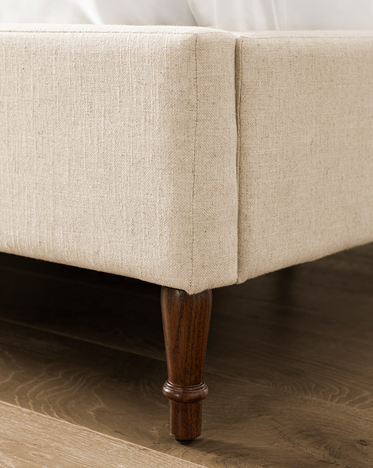 Close-up of the Willoughby Bed’s corner shows its beige linen upholstery and dark English Oak wood legs resting on a hardwood floor. White bedding is partially visible atop the bed frame, by Vendixs