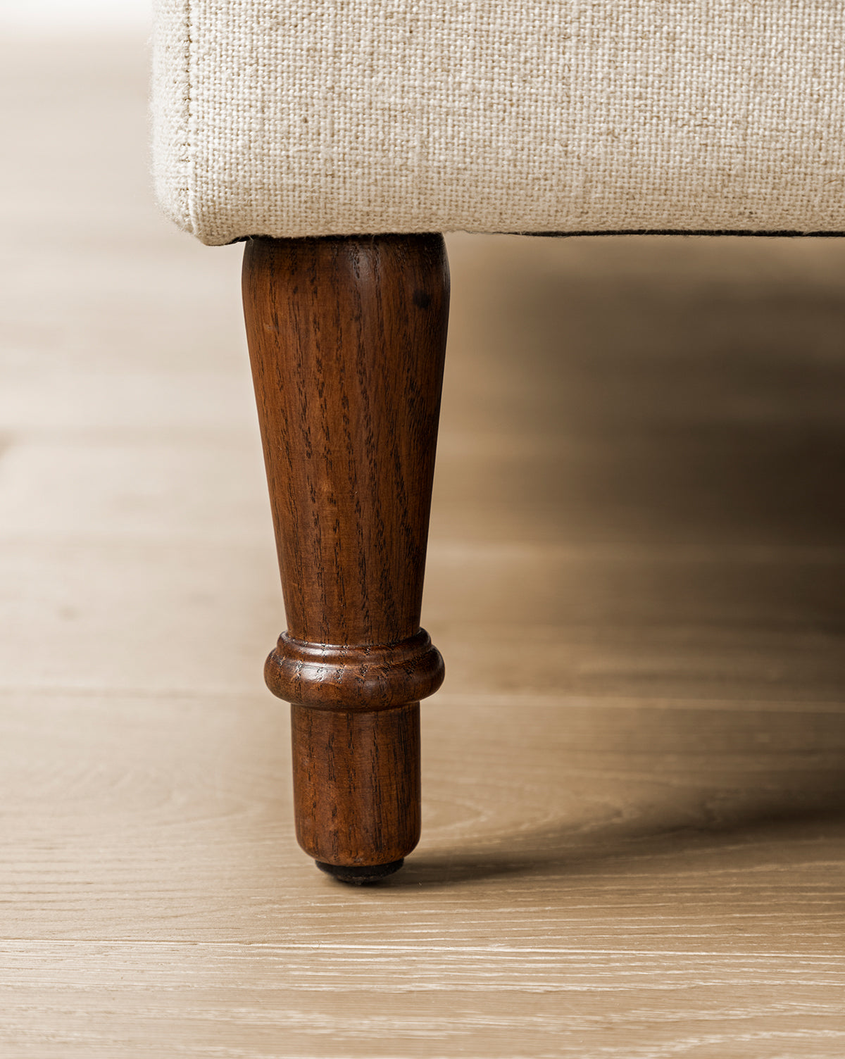 Close-up of an English Oak wood leg on the Willoughby Bed's beige linen upholstery, shown resting on a light wooden floor, by Vendixs