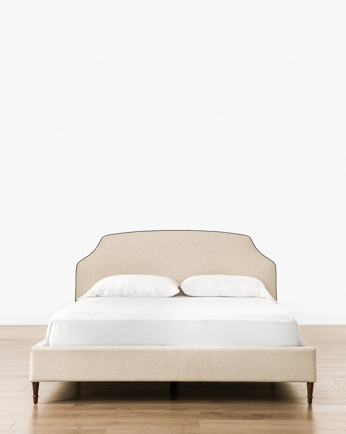 A beige Willoughby Bed with linen upholstery and a tall, curved headboard stands on elegant English Oak legs. It’s dressed in white sheets and pillows and set on a light wood floor against a plain white background, by Vendixs