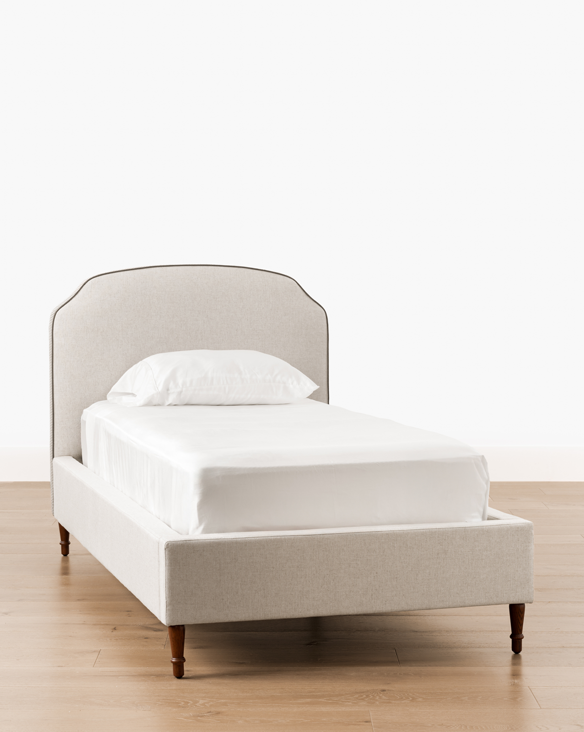 The Willoughby Bed features a light gray linen upholstered headboard and frame, white bedding, and English Oak legs, set on a light wood floor with a plain white background, by McGee & Co.