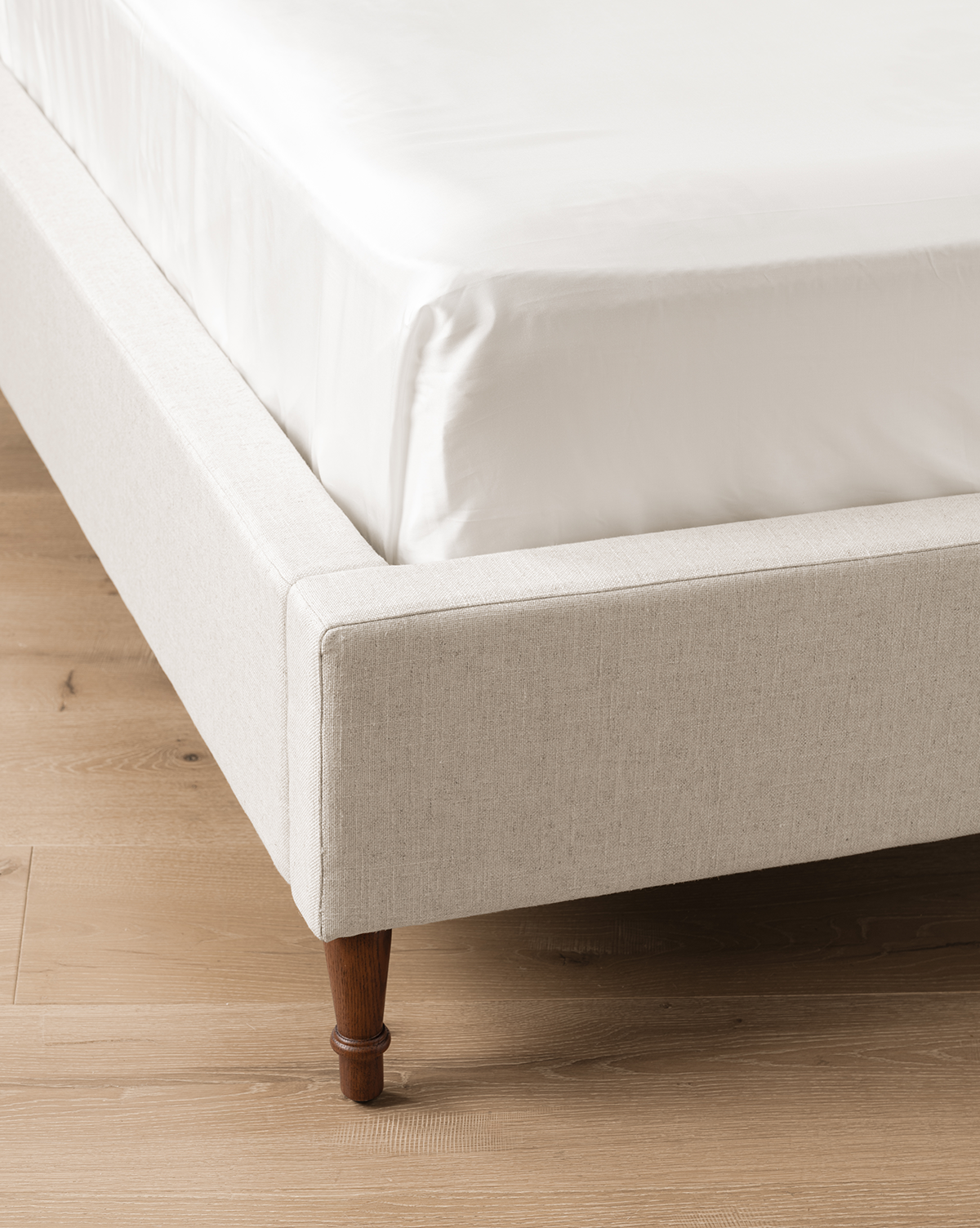 A close-up of the corner of the beige linen Willoughby Bed with English Oak legs, white mattress and bedding, set on a light wood floor, by Vendixs