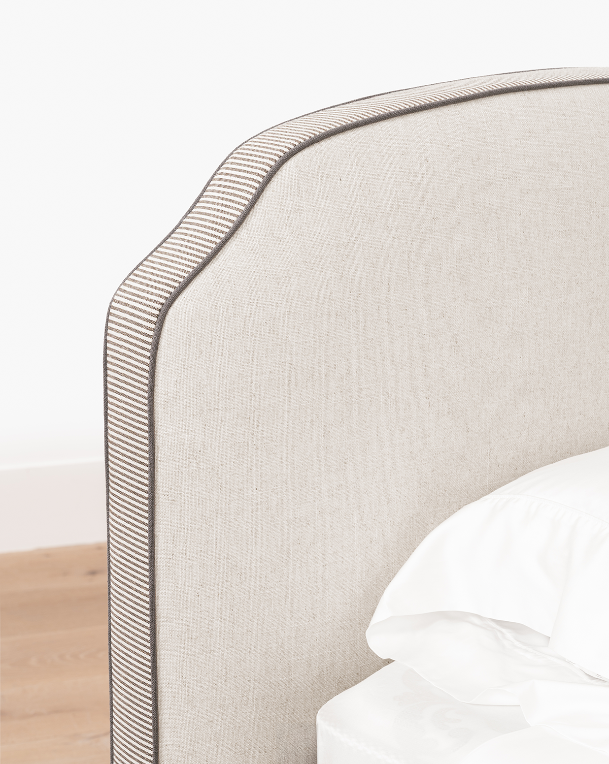 A close-up of the Willoughby Bed’s beige linen upholstered headboard, featuring a curved top and dark piping trim, shown beside white sheets and a light wood floor, by Vendixs