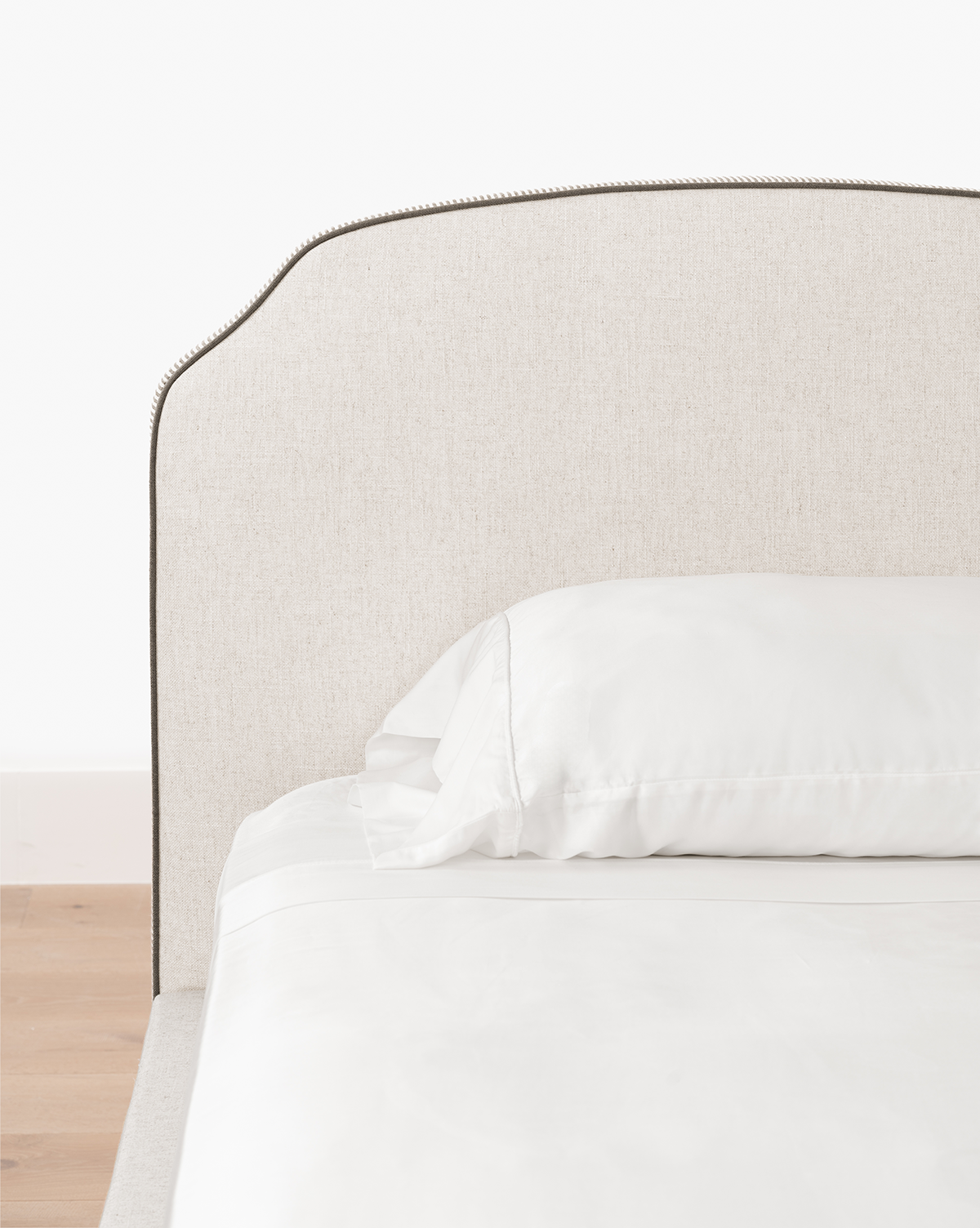 The Willoughby Bed features linen upholstery, a light beige headboard, white pillows and bedding, elegant English Oak legs, set against a white wall and light wood floor, by Vendixs
