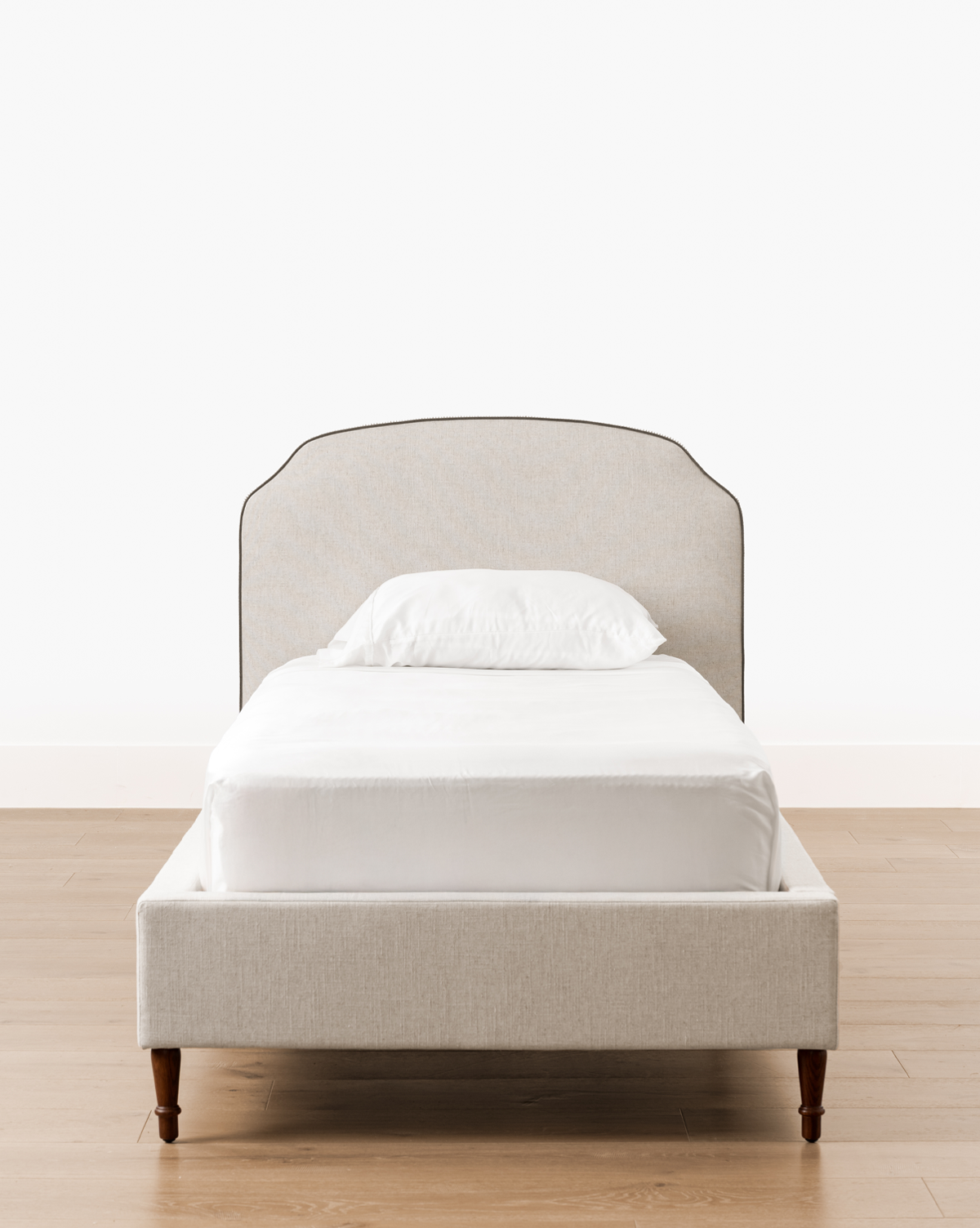A Willoughby Bed with light beige linen upholstery, English Oak legs, white bedding, and a single pillow stands on a wooden floor against a plain white wall, by Vendixs